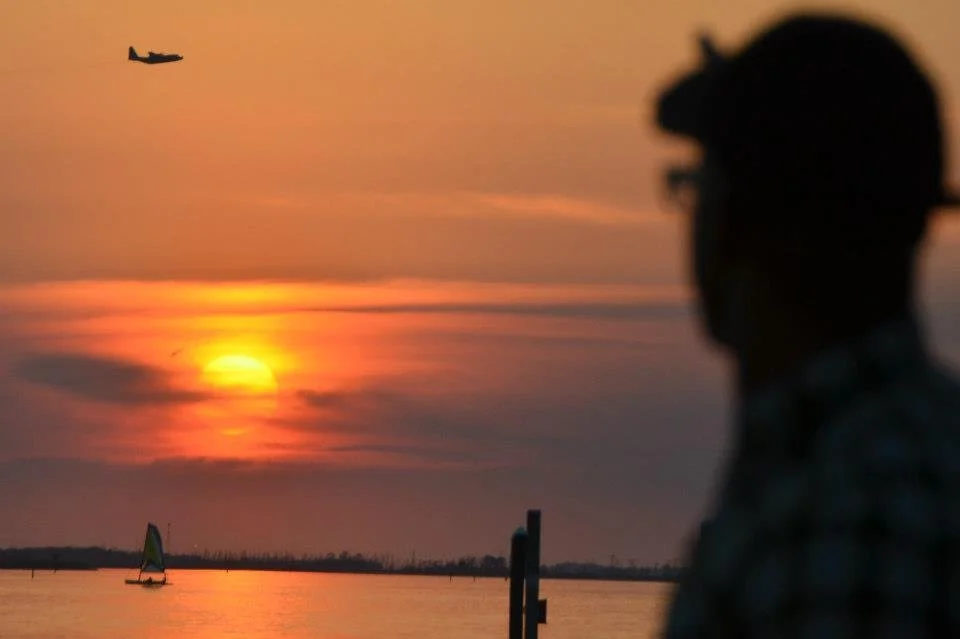 The author, blurry and silhouetted in the foreground, points to a distant aircraft flying low in the background. There is a low sunset beyond and a sailboat on calm water in the midground.