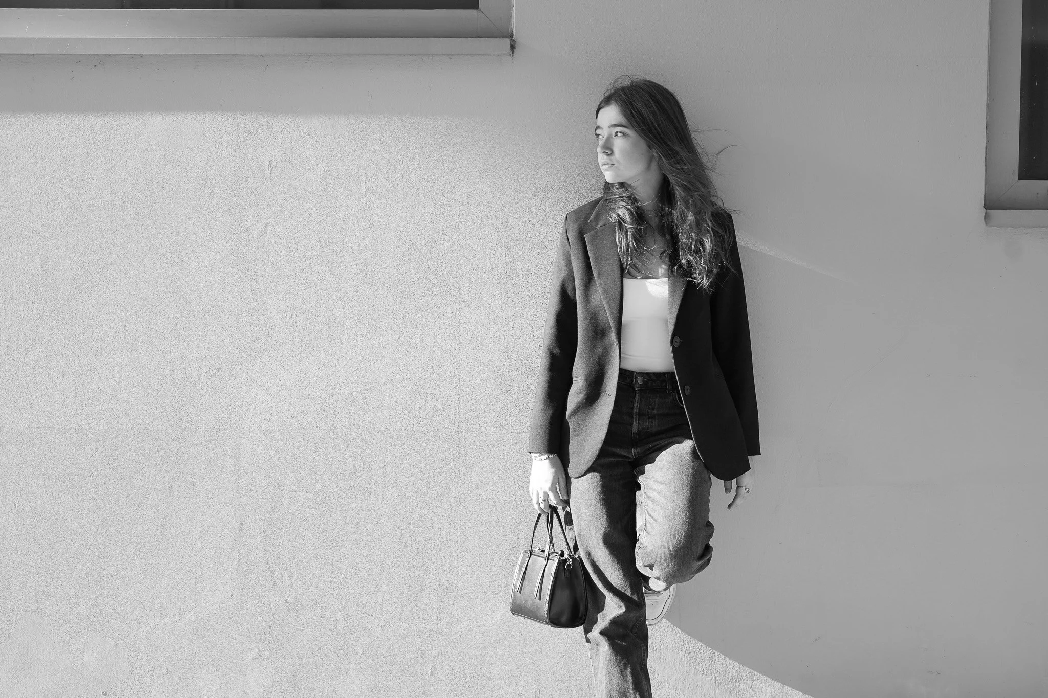Une jeune femme debout contre un mur blanc, portant une veste noire, un t-shirt blanc, un jean noir, et tenant un sac à main, regardant vers la droite.