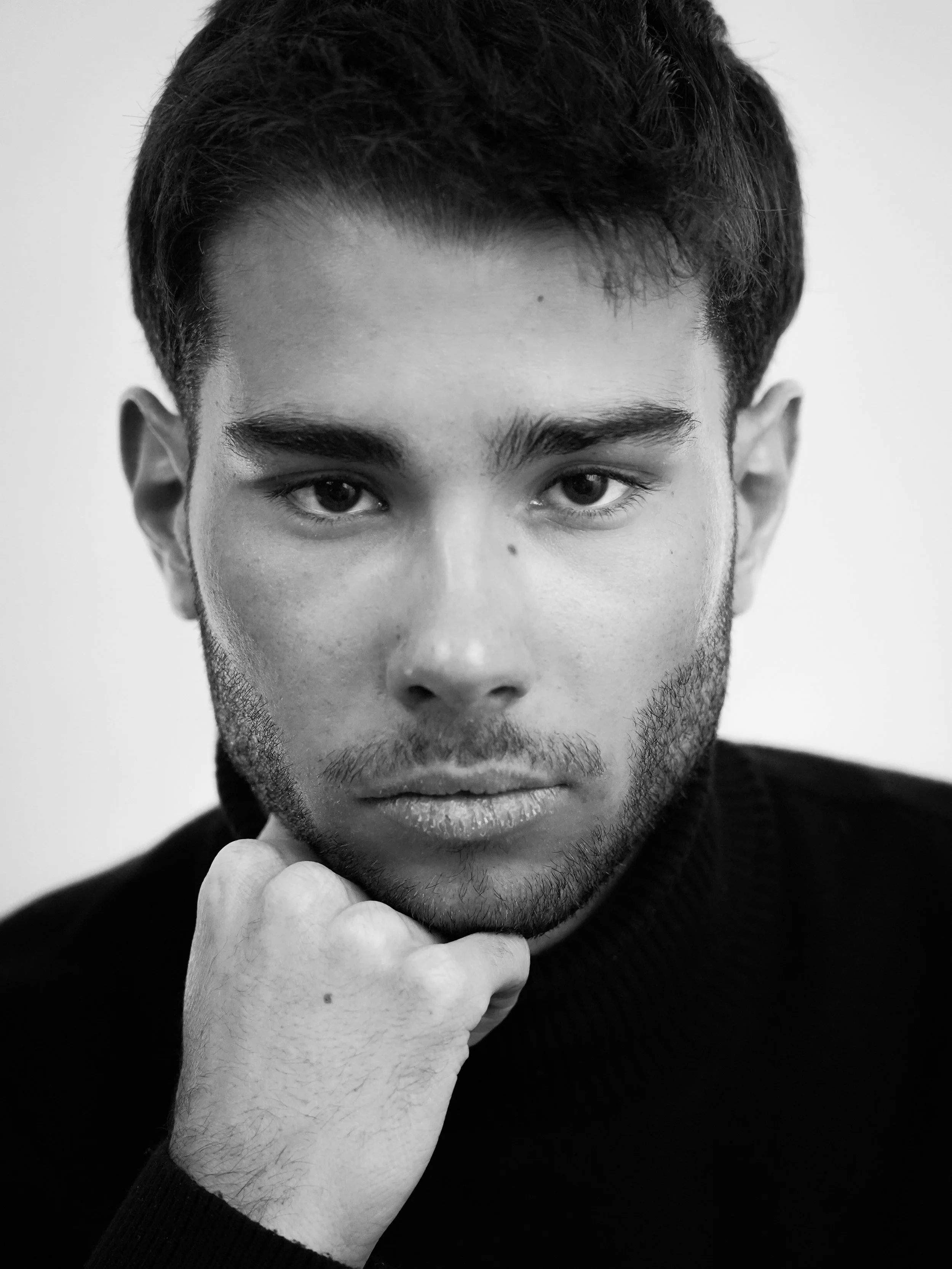 Un portrait en noir et blanc d'un jeune homme regardant directement l'appareil, avec le menton posé sur sa main, portant un pull noir.