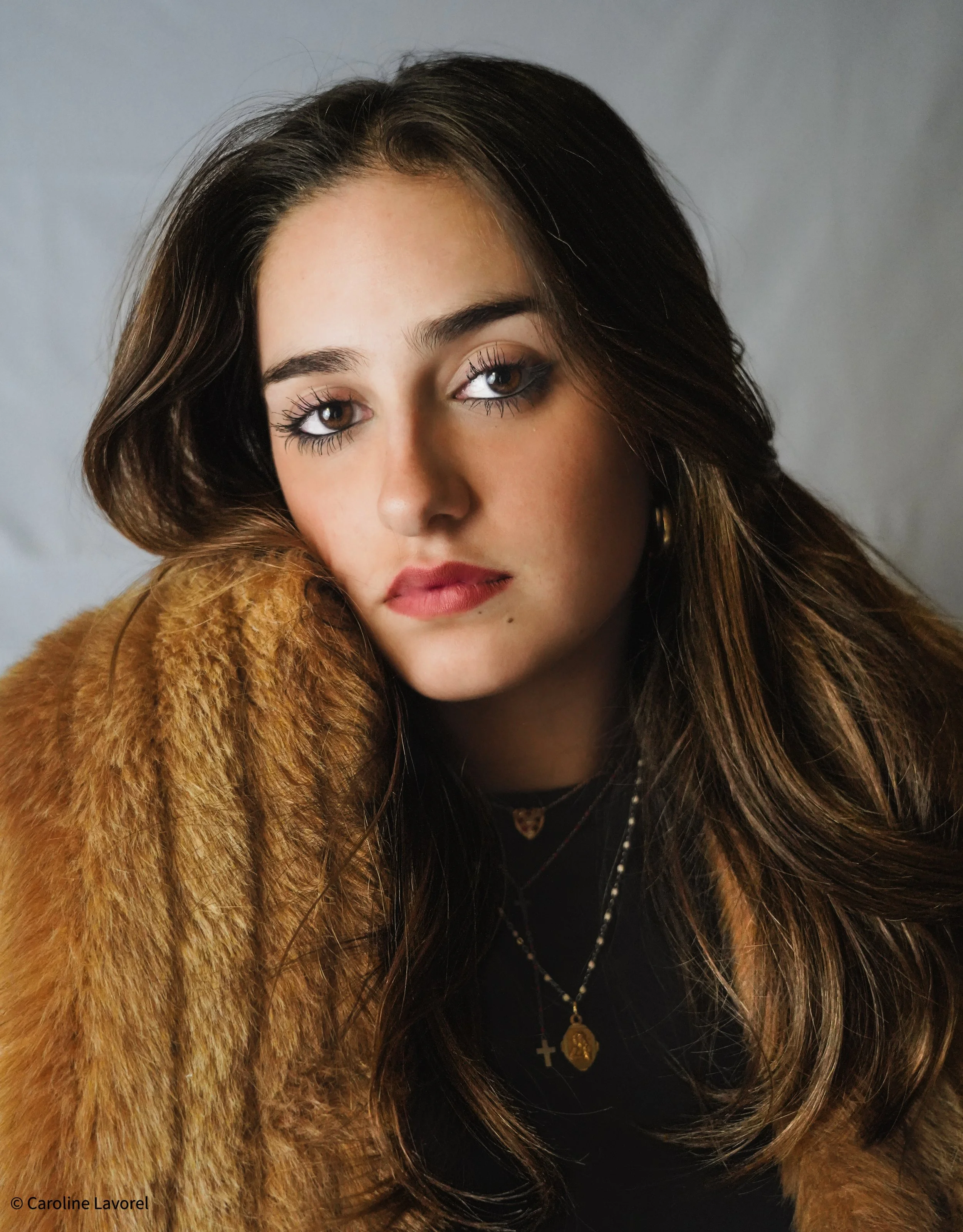 Portrait d'une jeune femme avec des cheveux bruns, portant un manteau en fourrure, des bijoux, et un maquillage léger, regardant vers l'objectif. En arrière-plan gris clair.