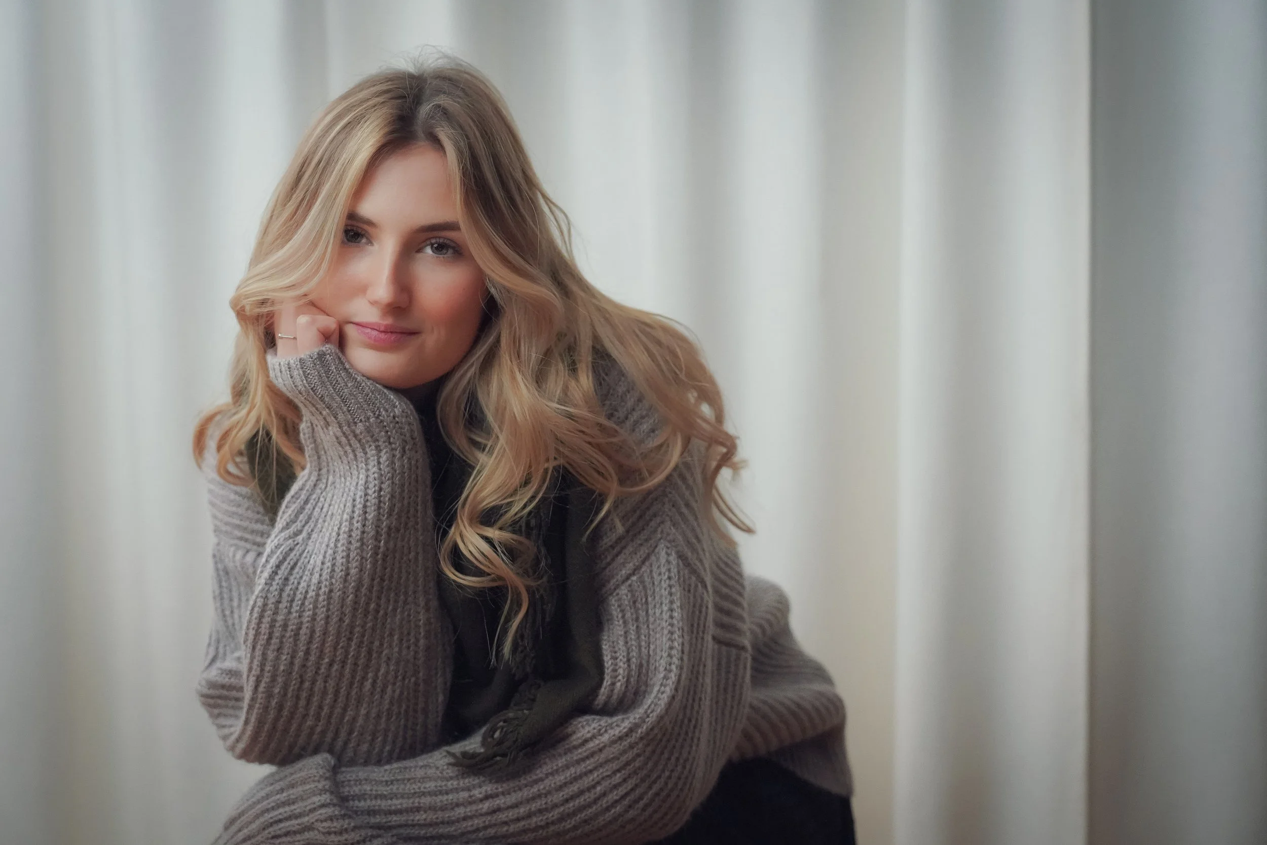 Jeune femme aux cheveux blonds bouclés, portant un pull gris, posant avec sa main sous son menton dans un intérieur lumineux.