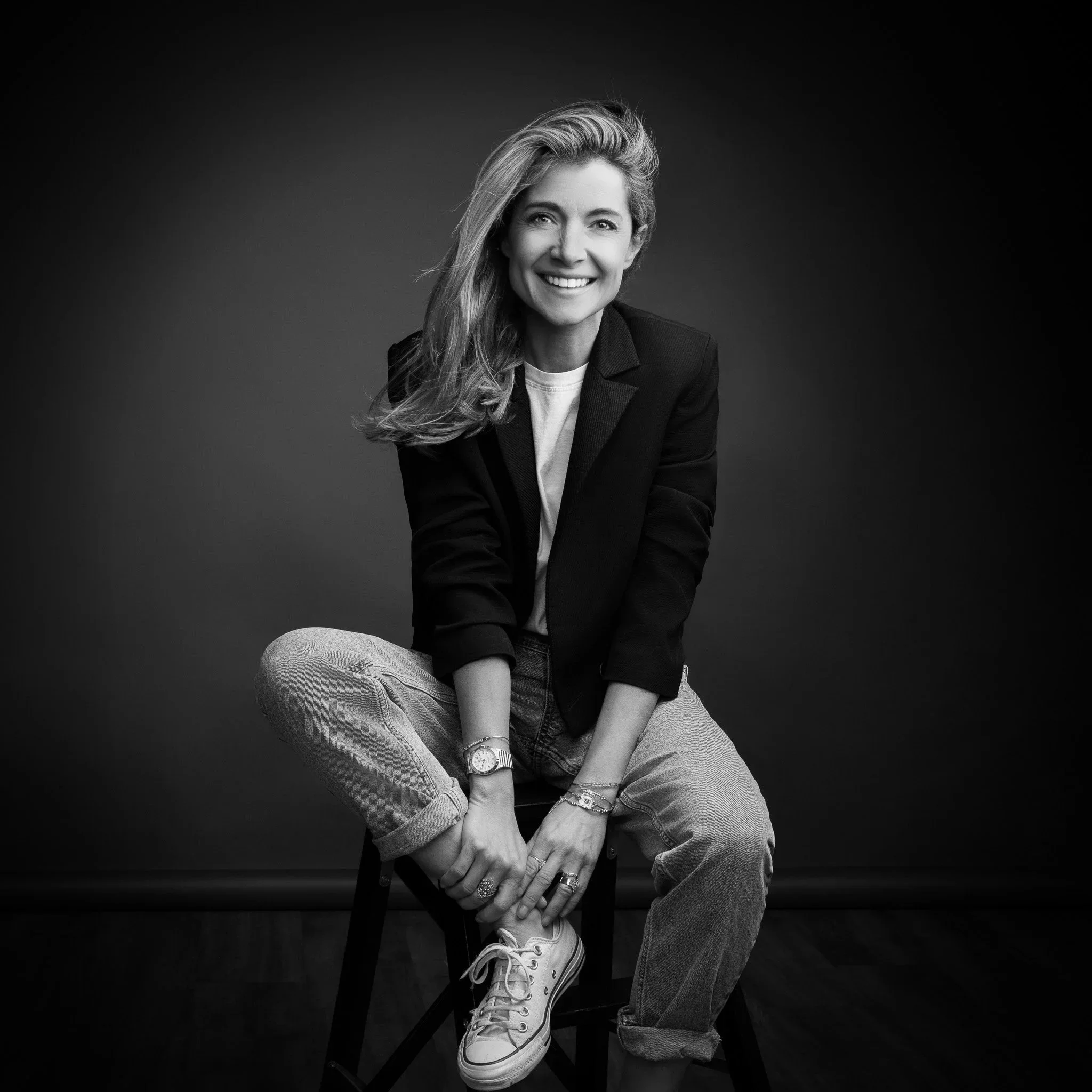 Portrait en noir et blanc d'une femme assise sur une chaise, souriante, portant un blazer noir, un t-shirt blanc, un jean et des Converse blanches, avec des bijoux et une montre.
