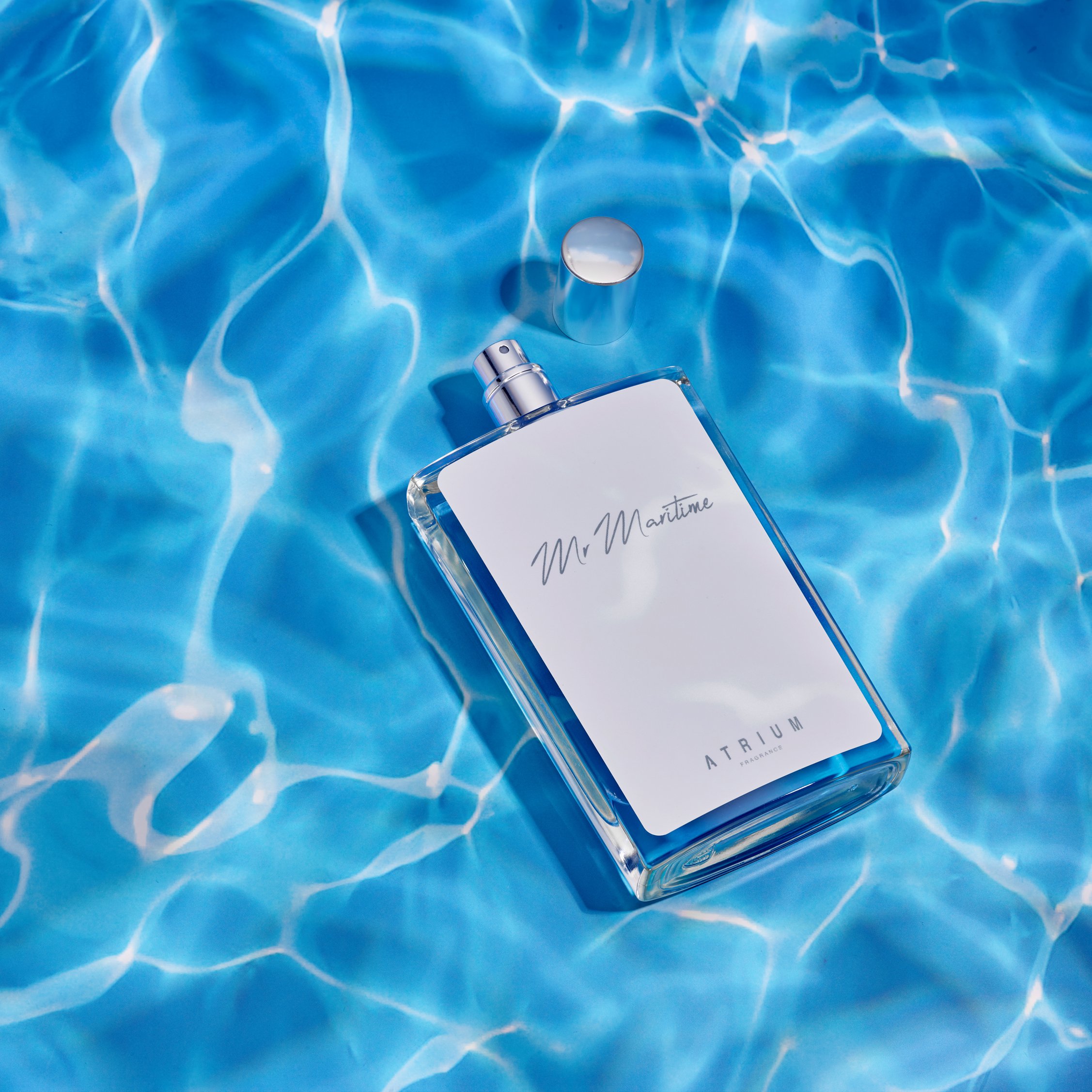 A clear glass perfume bottle with a silver cap and the label 'Mr. Maritime' on a light blue rippling water background.