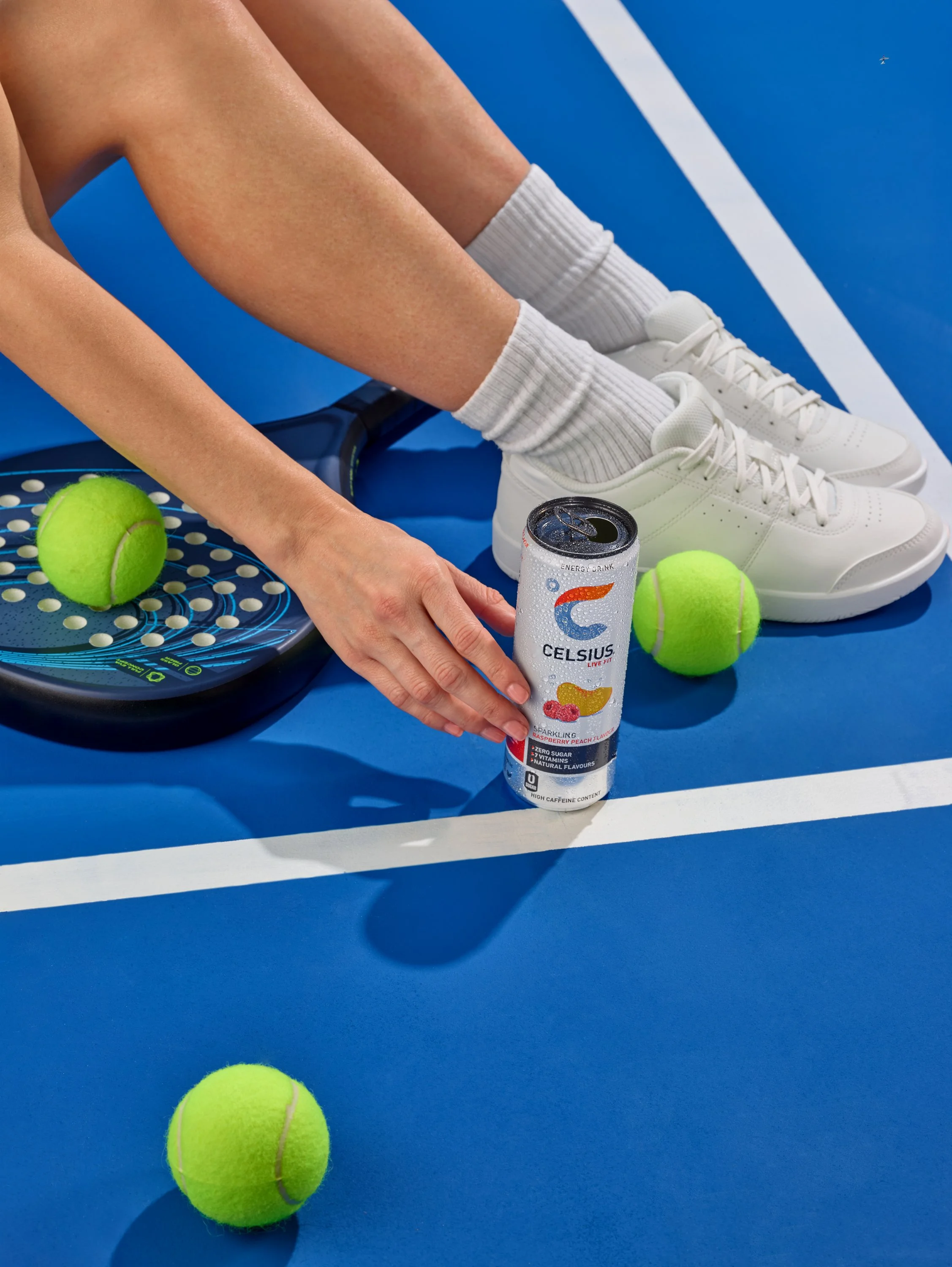 model sits on paddle court surround by tennis balls reaching for drinks can