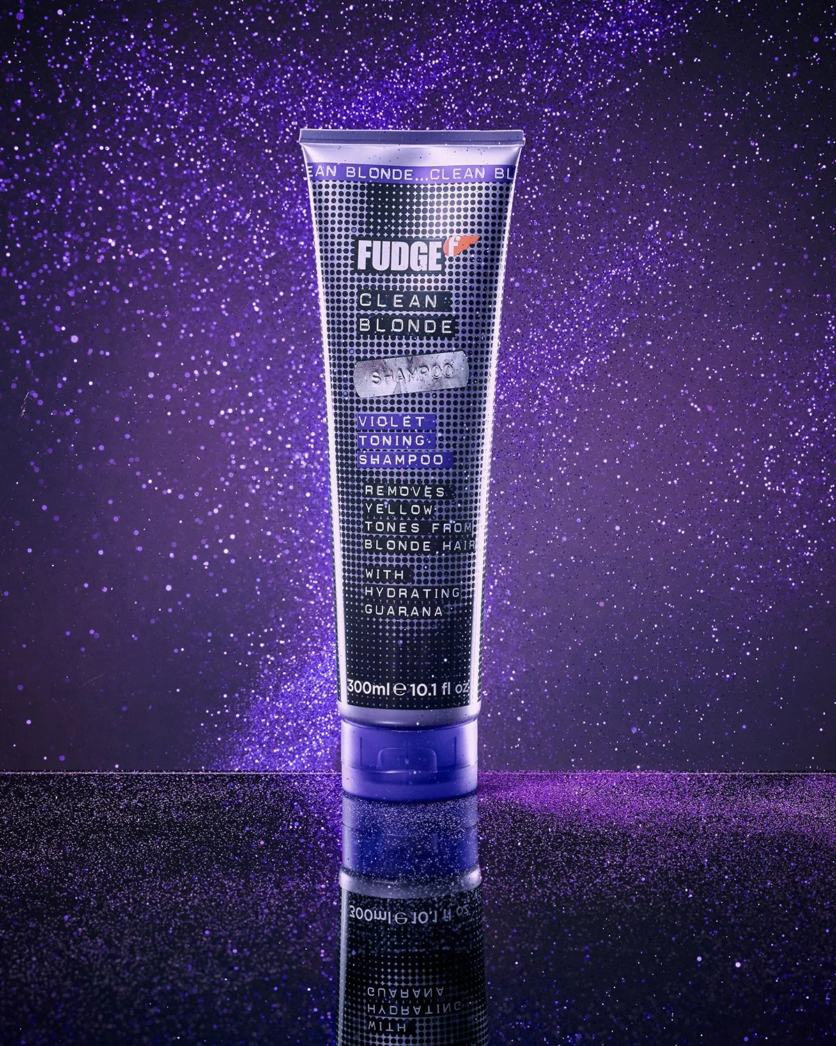A tube of Fudge Clean Blonde Violet Toning Shampoo standing upright on a glittery purple surface with a sparkling purple background.