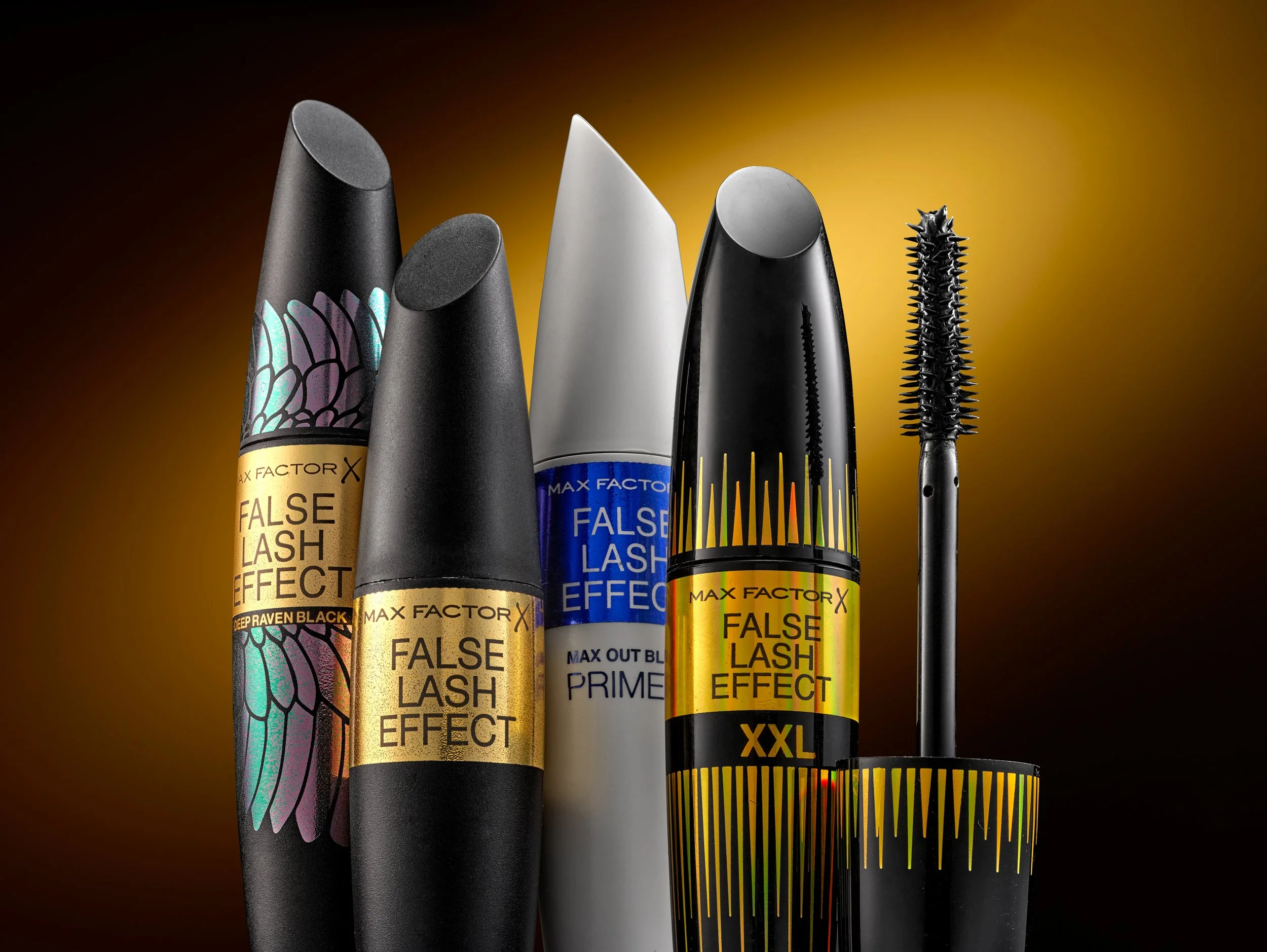 Four Max Factor False Lash Effect mascaras in different colors and designs with a mascara brush next to them, set against a dark gradient background.
