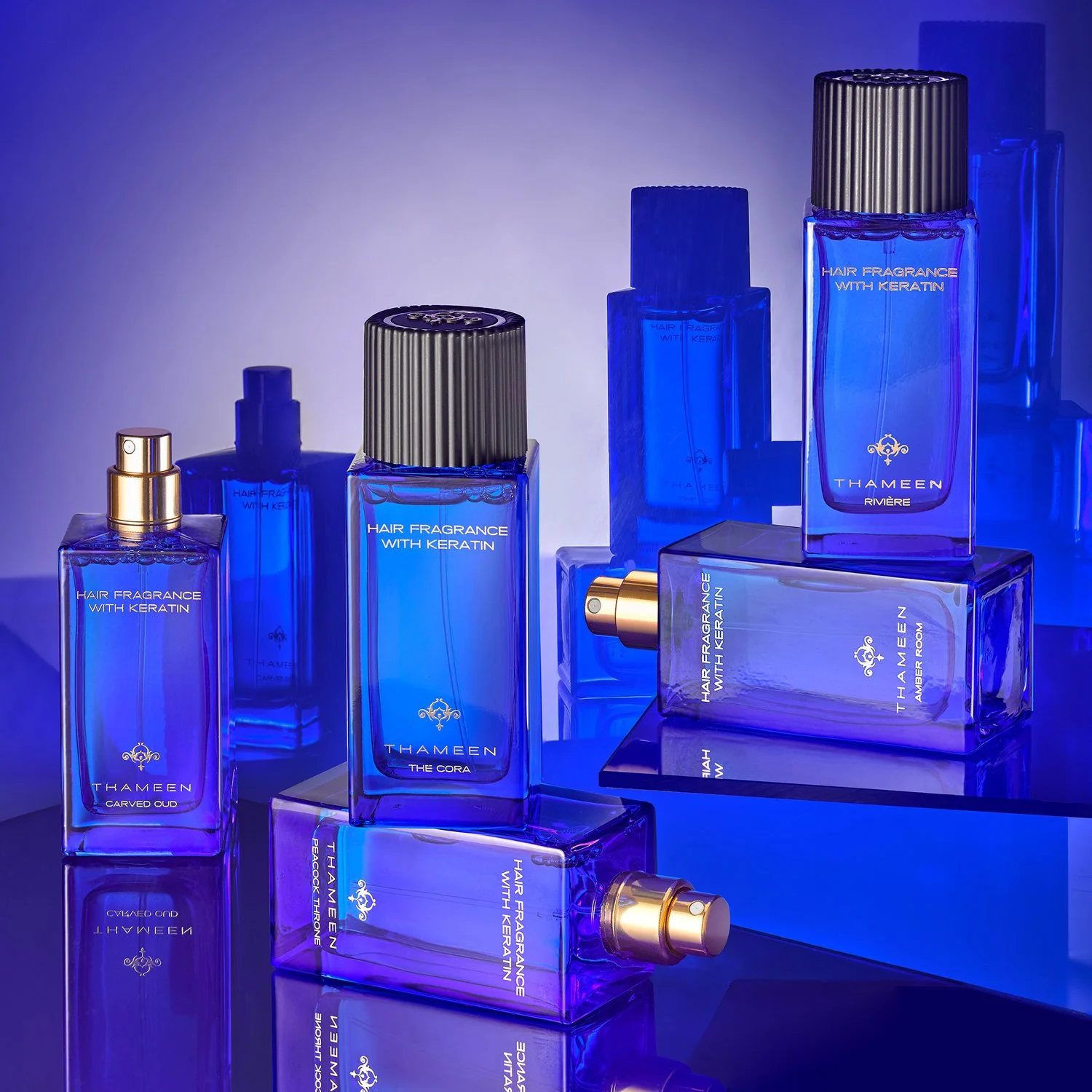 Multiple blue perfume bottles with gold caps, labeled with names such as 'Carved Oud', 'The Cora', and 'Amber Room'. The bottles are arranged on reflective surfaces with blue lighting and background.