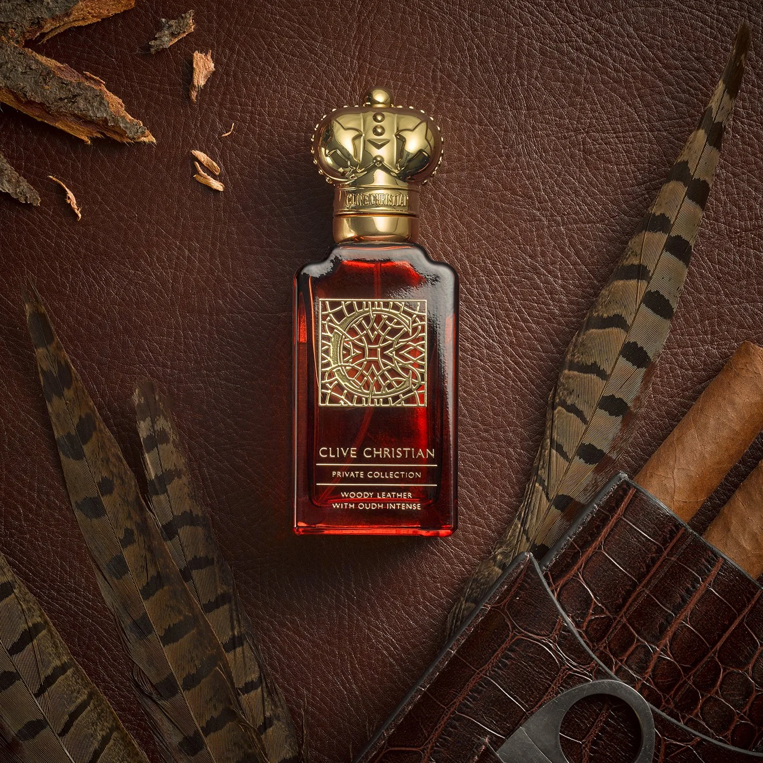 A bottle of Clive Christian private collection woody leather perfume with a gold crown-shaped cap, placed on a dark brown leather surface, surrounded by feathers, a rolled cigar, a brown leather item, and a metallic item with circular opening.