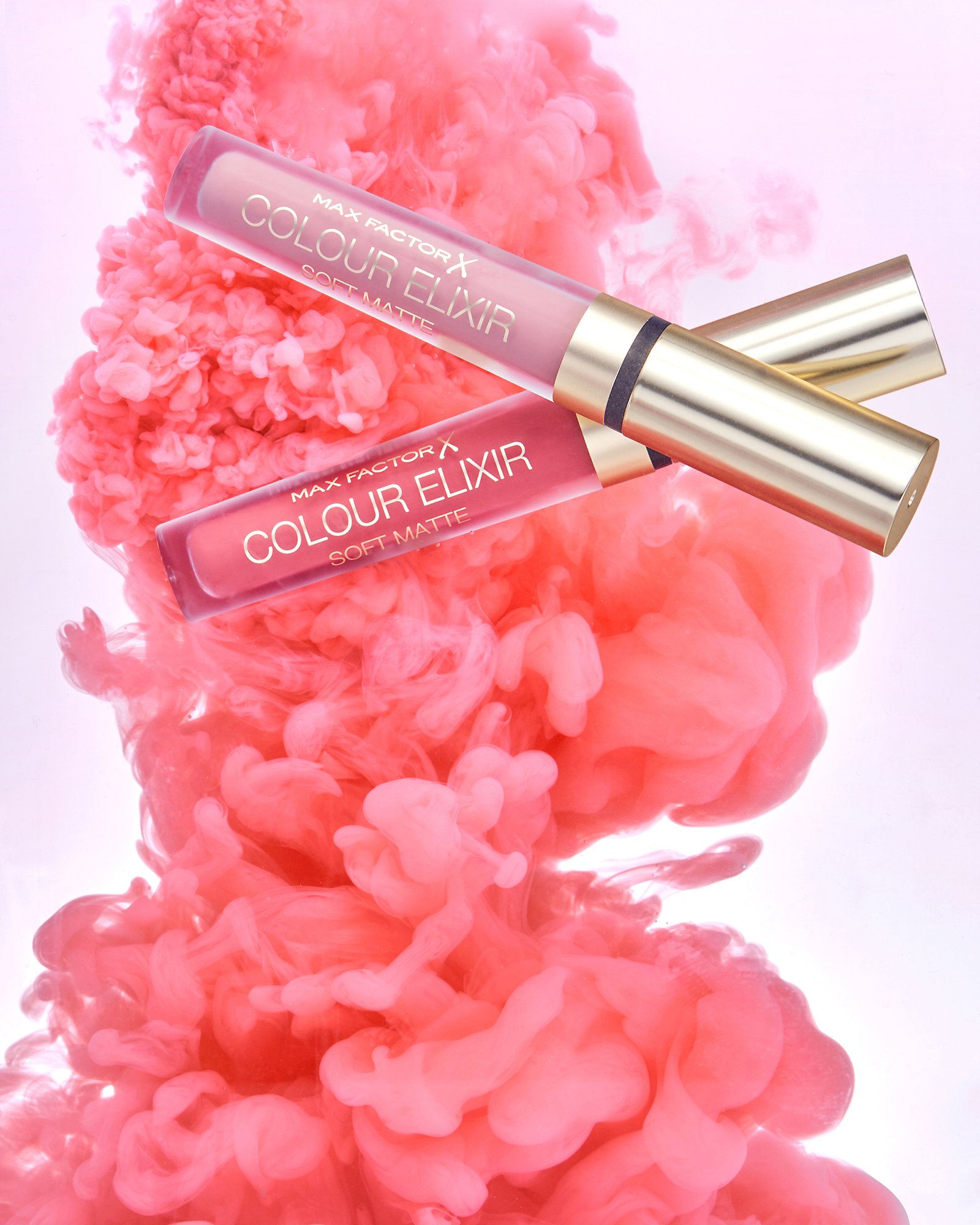 Two tubes of Max Factor Colour Elixir lipstick, one soft matte and one glossy, placed on a pink and peach-colored ink cloud background.