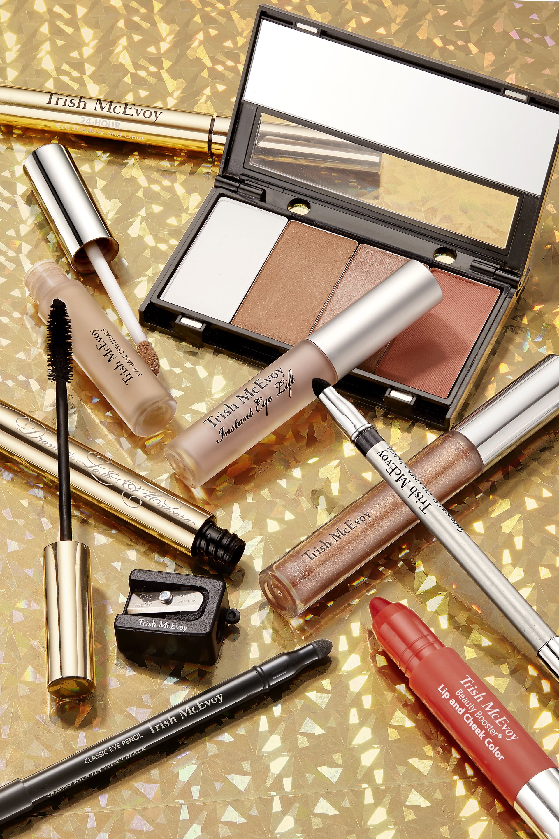 Makeup products, including an eyeshadow palette and various eyeliners, are arranged on a sparkling gold surface.