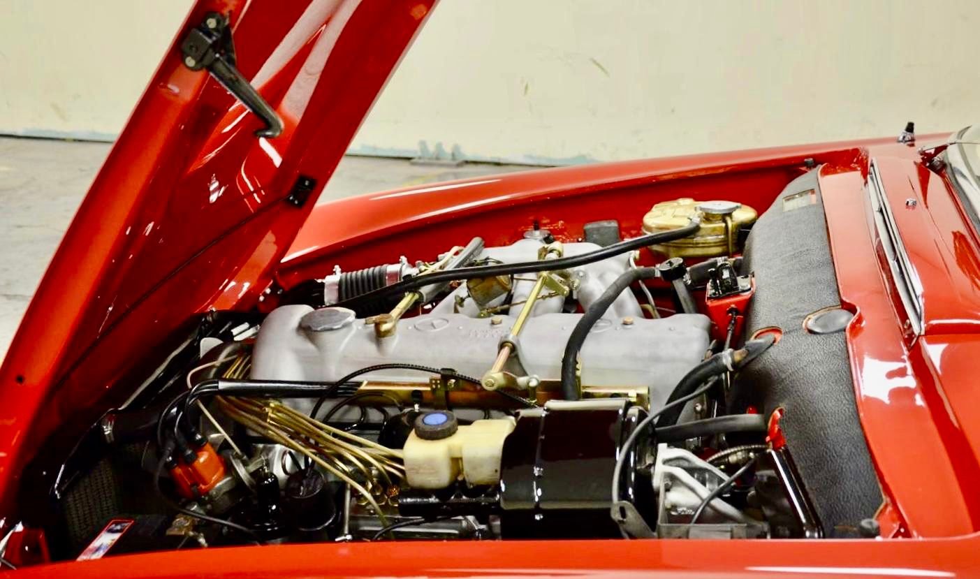 Close-up of a vintage red sports car engine with various metallic and black components, hoses, wires, and a black fabric-covered dashboard at the front.