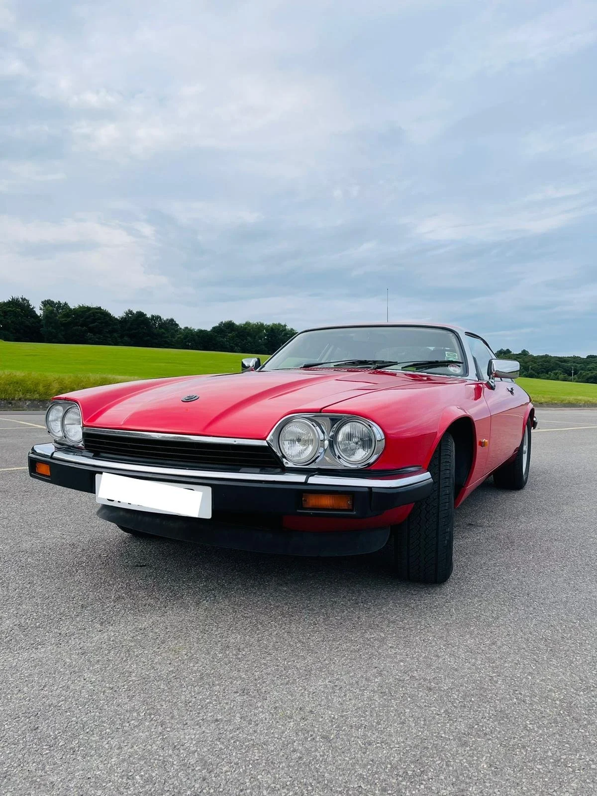 Red vintage sports car with black trim parked on a paved surface, with green grass and trees in the background under a partly cloudy sky.