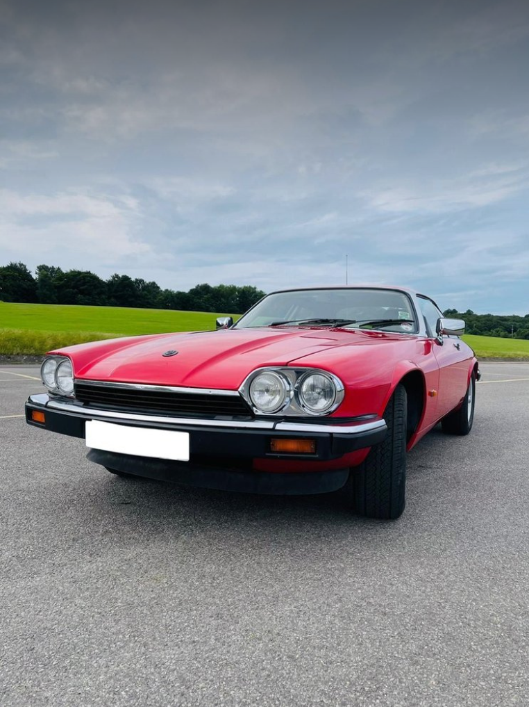 A red vintage sports car parked on a paved road with green fields and trees in the background, under a cloudy sky.