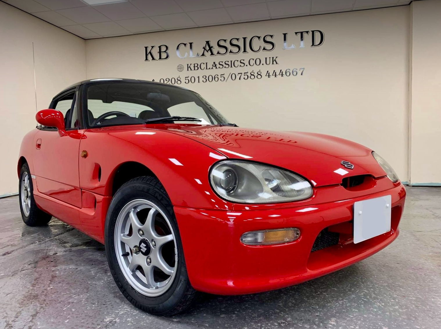 A red Suzuki sports car parked indoors with a black roof, in front of a wall with black text that reads "KB Classics Ltd" and contact information.