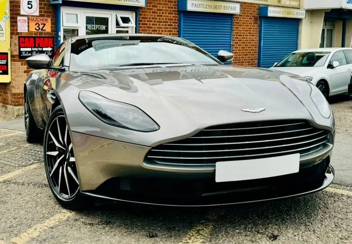 A silver Aston Martin sports car parked in front of a building with multiple signboards. The building has brick and white walls, blue shutters, and various signs advertising car repairs and bodywork services. There is also a white car and a black car