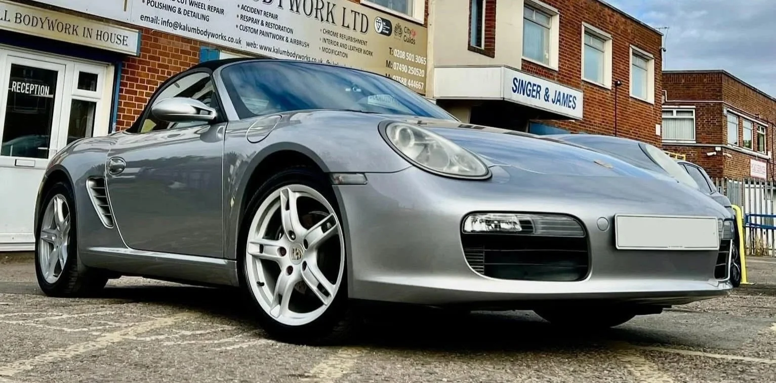 A silver Porsche convertible sports car parked in front of a commercial building with signage for Kalum Bodywork Ltd, a tattoo studio, and a sign for Singer & James, with brick and beige facades, under a partly cloudy sky.