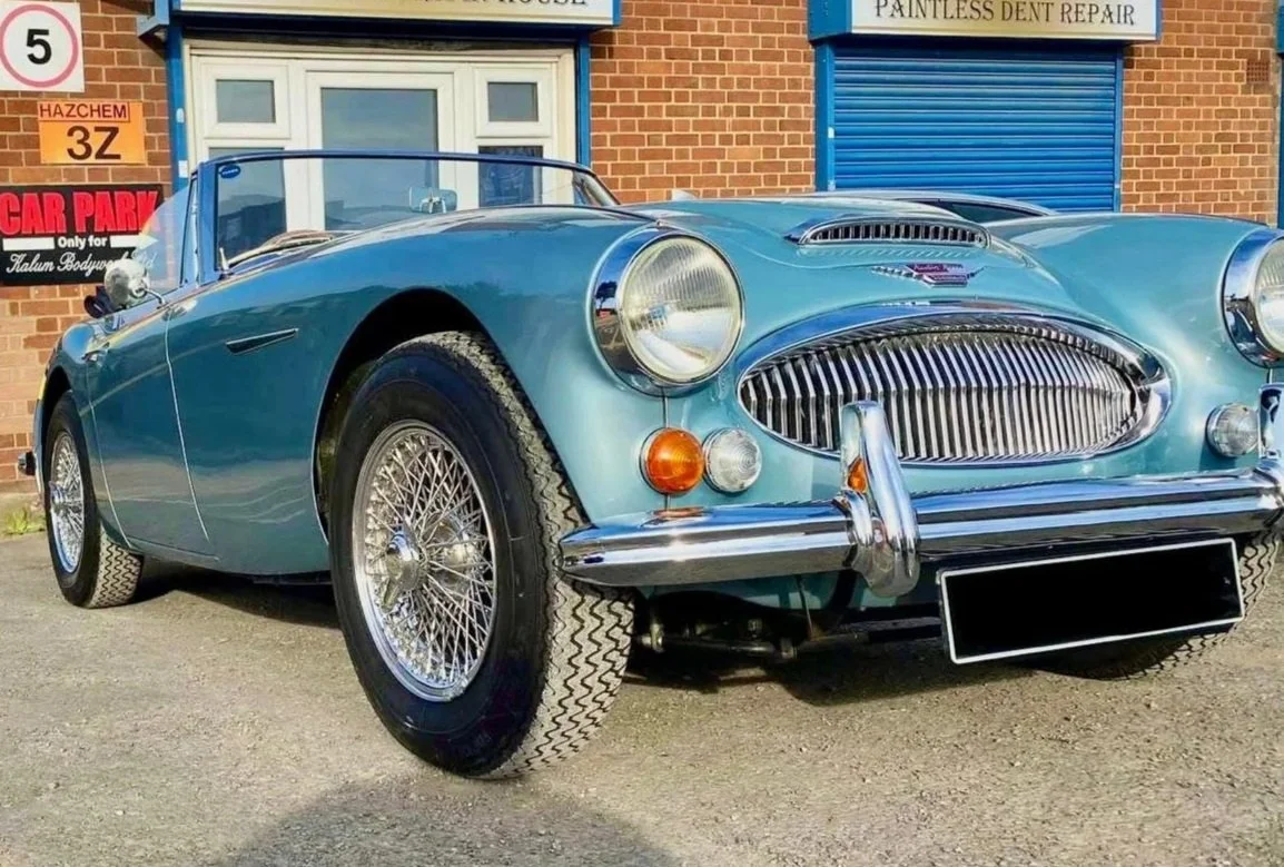 A vintage blue convertible sports car parked outside a brick building with signs for a bodywork shop, featuring chrome accents, wire wheels, and classic design elements.