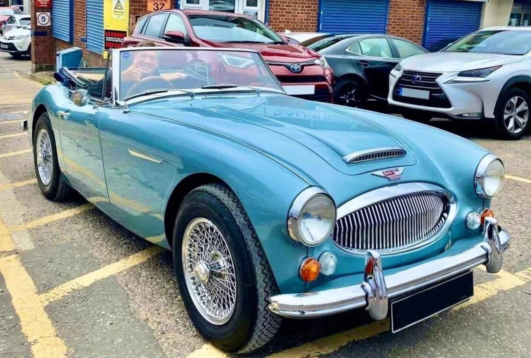 A light blue vintage convertible car parked in a lot with other modern vehicles. The car has chrome wire wheels and a classic design, with a man smiling and giving a thumbs-up in the driver's seat. There are multiple business signs on a brick buildin