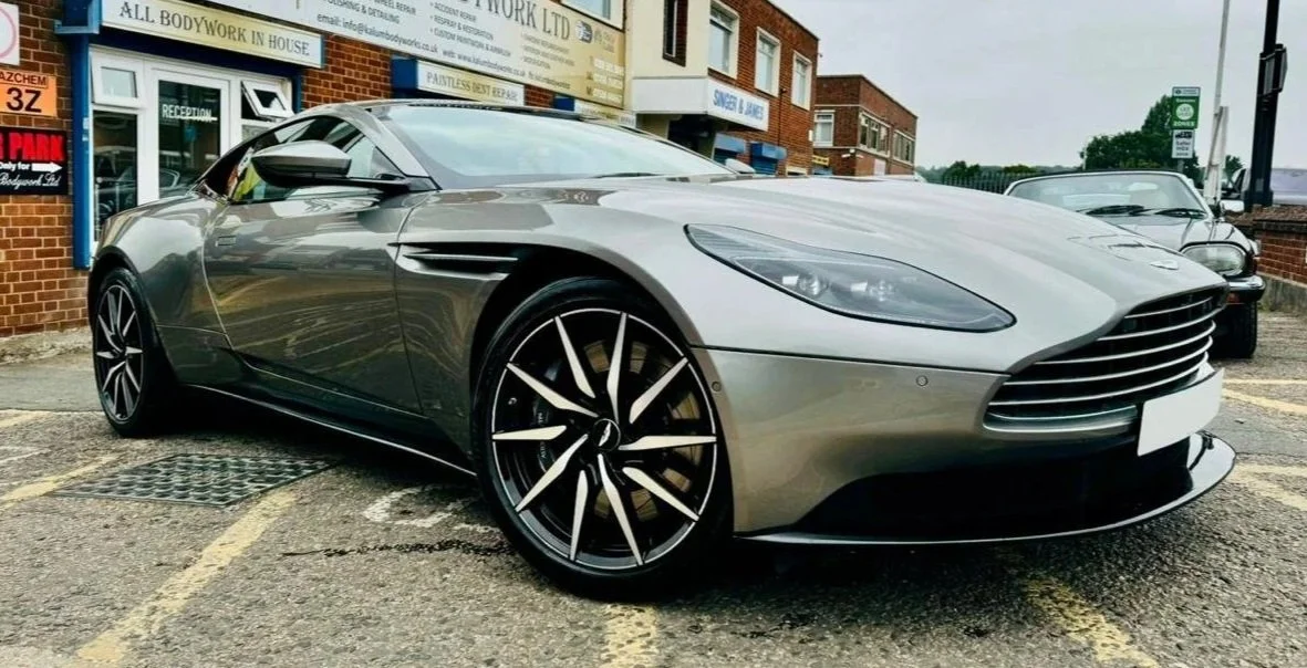 A silver luxury sports car parked outside a commercial building, with other vintage cars visible in the background.