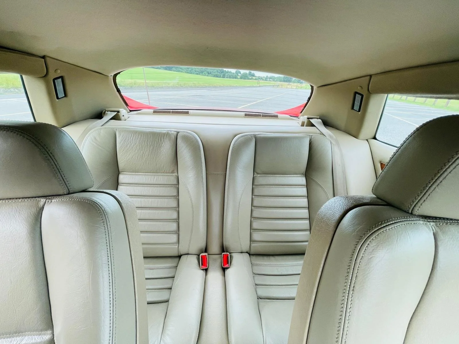 Interior view of a beige leather back seat of a car, with a view through the rear window of an empty parking lot and green landscape beyond.