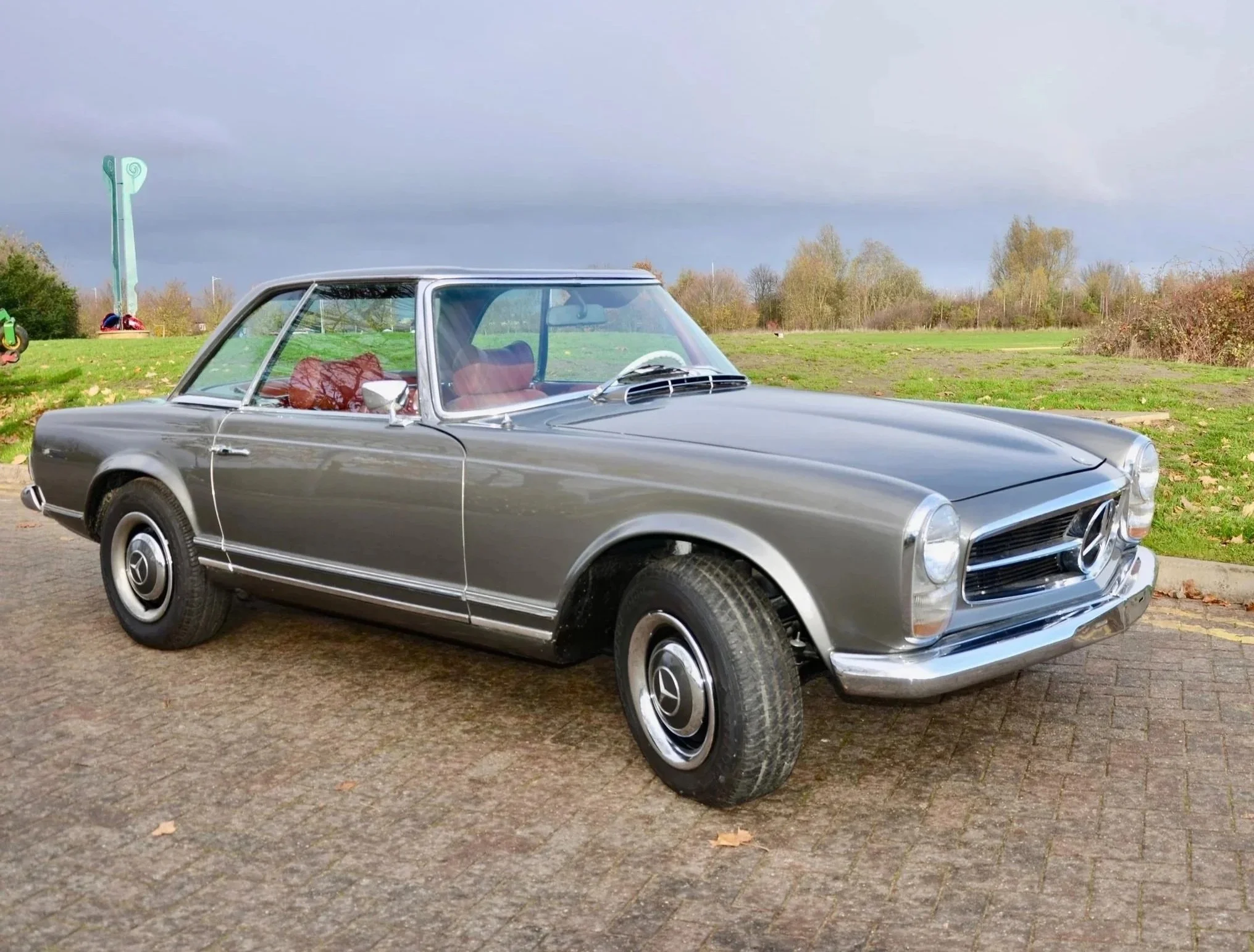 A vintage silver convertible car parked on a paved area with green grass and trees in the background.