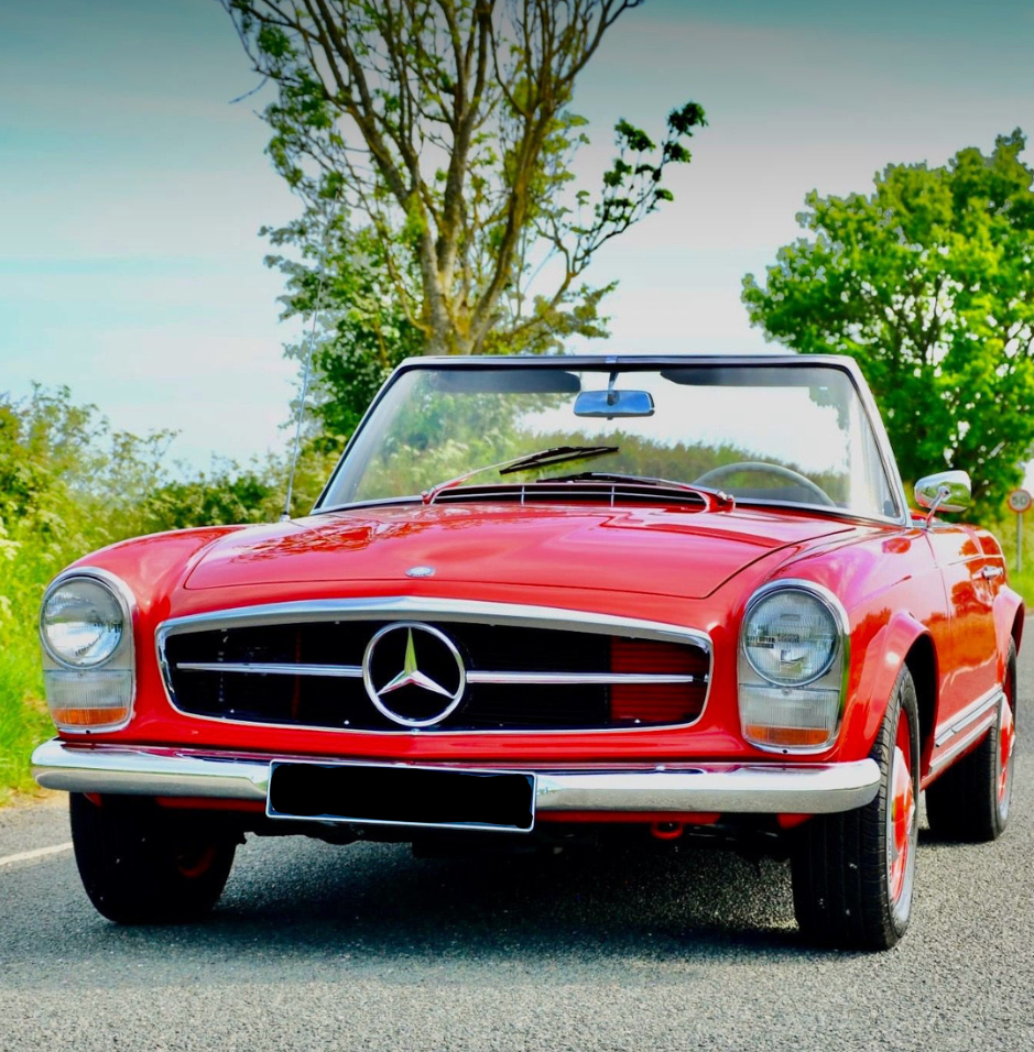Red vintage Mercedes-Benz convertible car parked on a rural road, with green trees and blue sky in the background.
