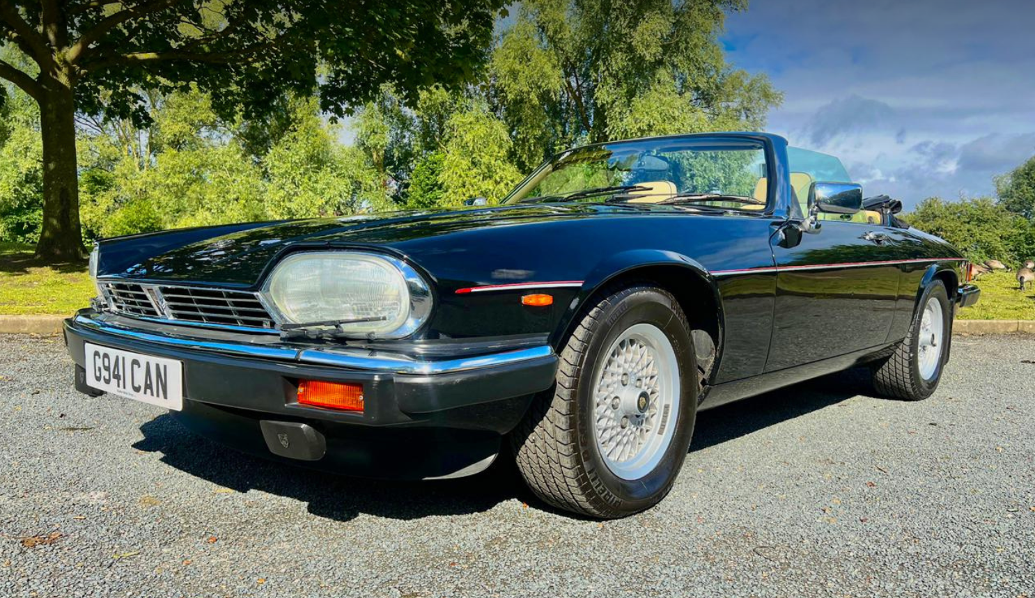 A classic black convertible car parked on a gravel surface with green trees and a blue sky in the background.