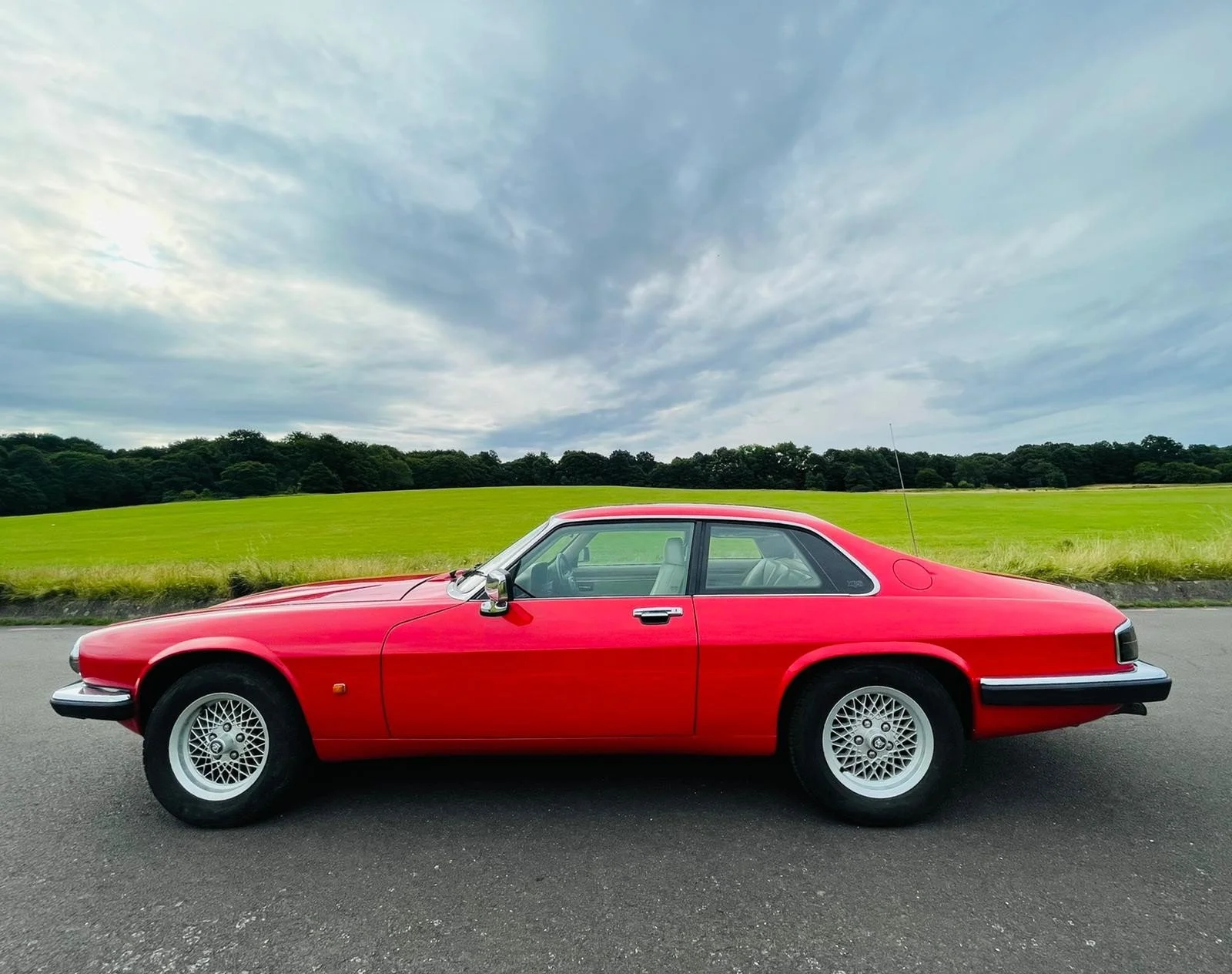 A vintage red car parked on a paved road with a lush green field and a cloudy sky in the background.