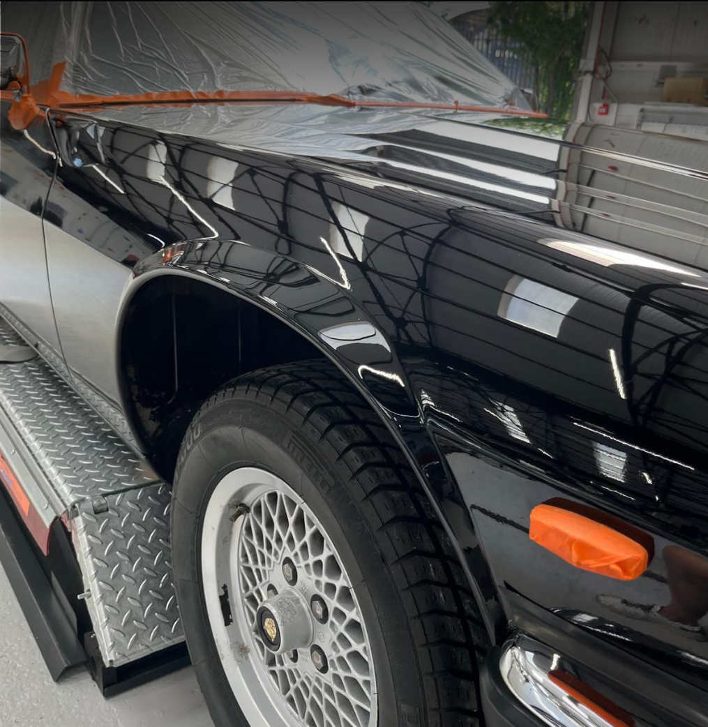 Close-up of a black vintage sports car with shiny paint, parked on a metallic platform in a garage or workshop, with orange trim on the side marker.