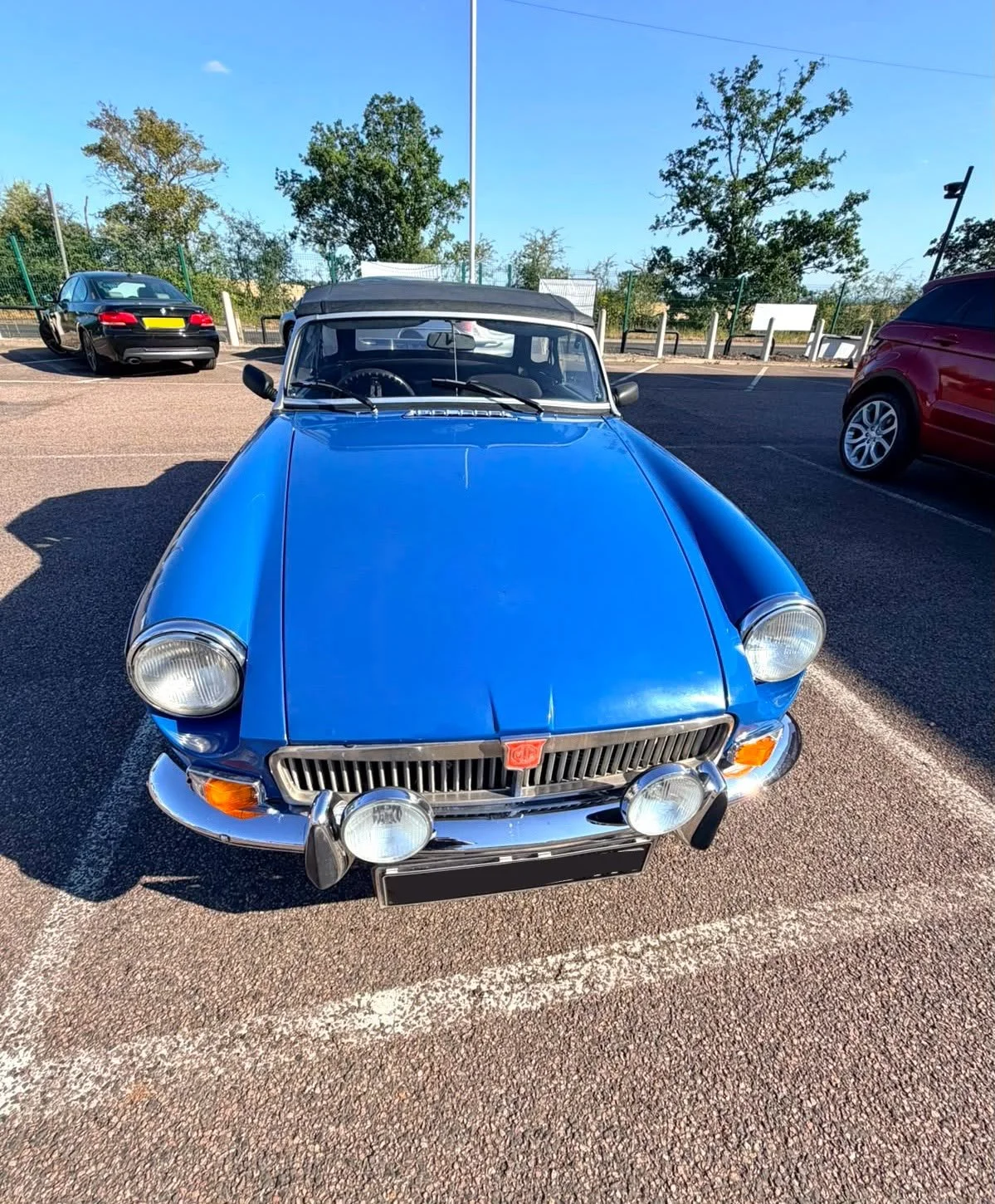 Front view of a vintage blue car parked in a parking lot, with other vehicles and trees in the background.