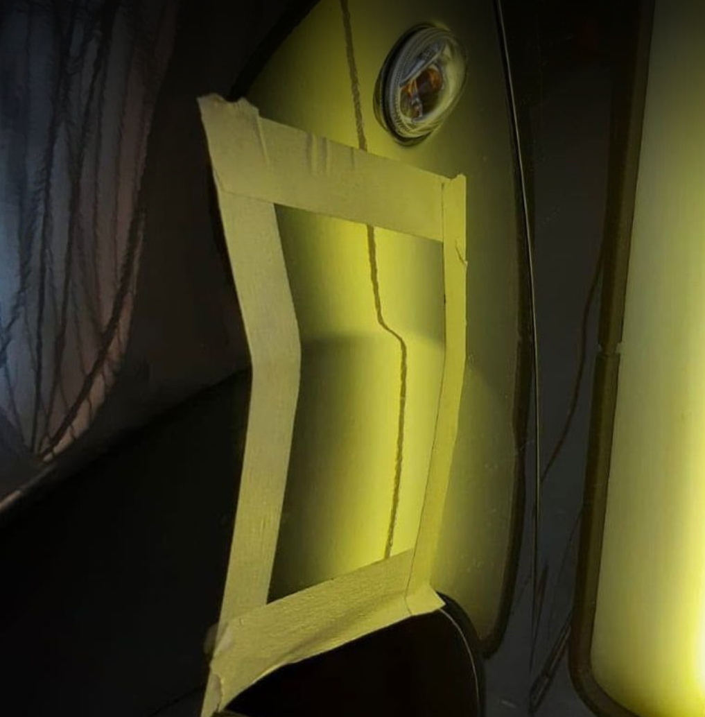 A black car with yellow masking tape forming a rectangular shape on its hood. There is a circular light or sensor on the car's roof near the window. The car appears to be parked indoors, possibly in a garage.