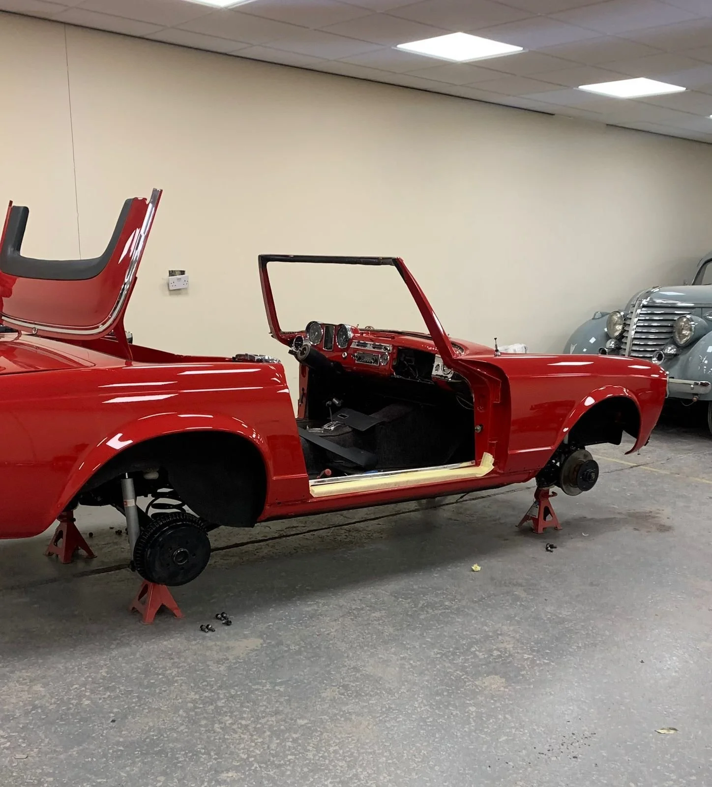 Red vintage car body with no wheels, artwork in progress or restoration, supported by jack stands in a garage with another classic car in the background.