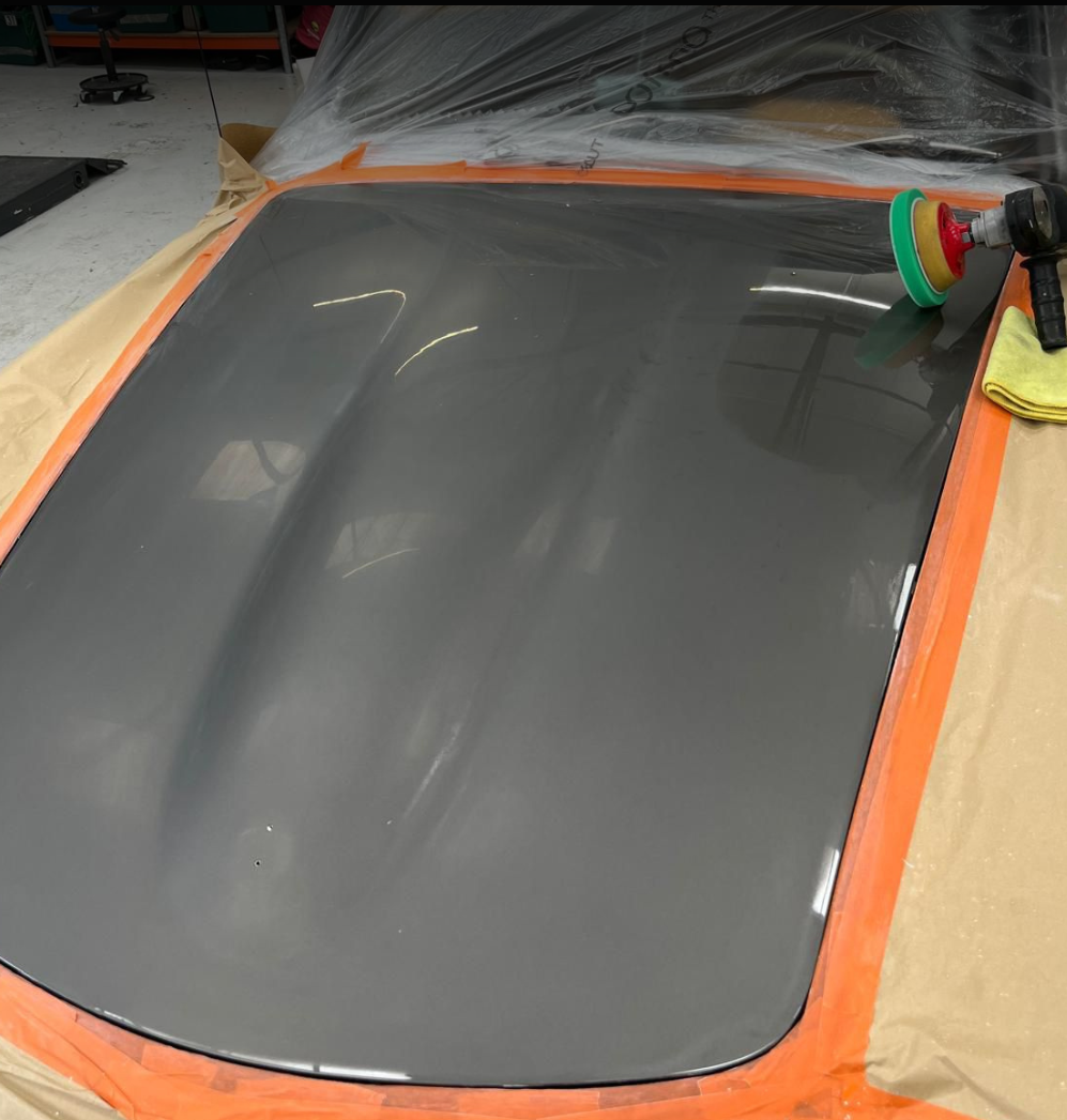 Close-up of a car hood being polished with a power buffer, surrounded by masking tape and protective paper in a workshop.