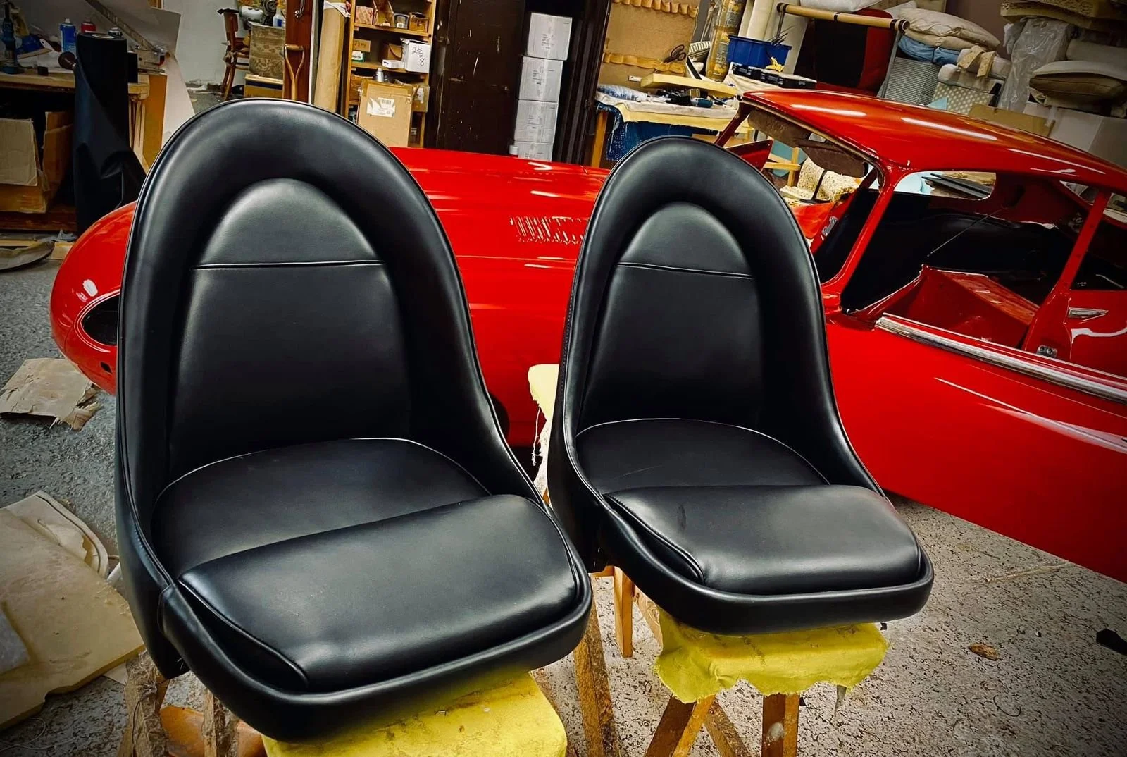 Two black leather car seats positioned on wooden stools in a workshop, with a red vintage car body in the background.