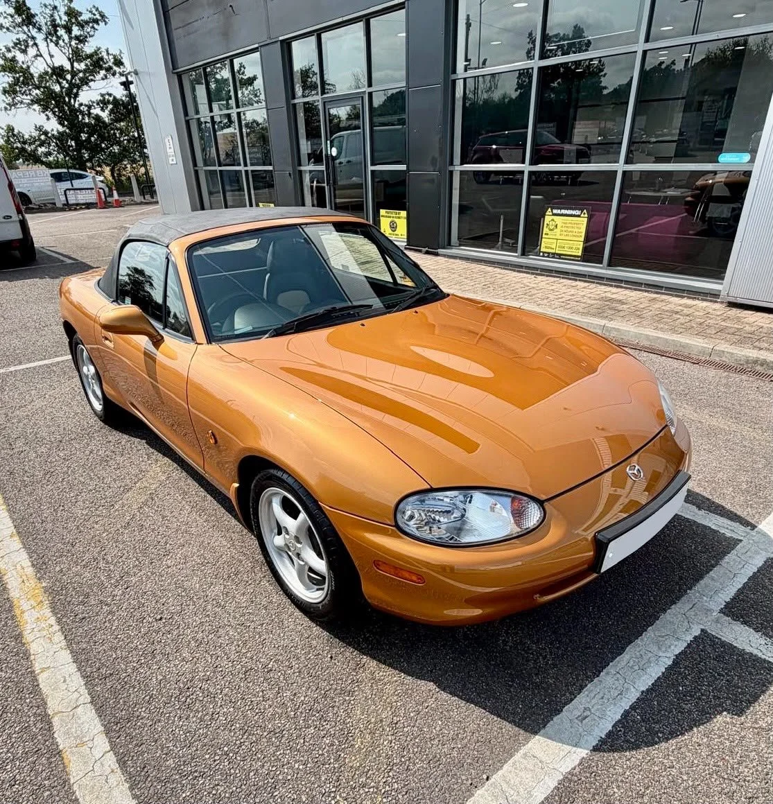 An orange Mazda MX-5 Miata convertible parked outside a commercial building with reflective glass windows.