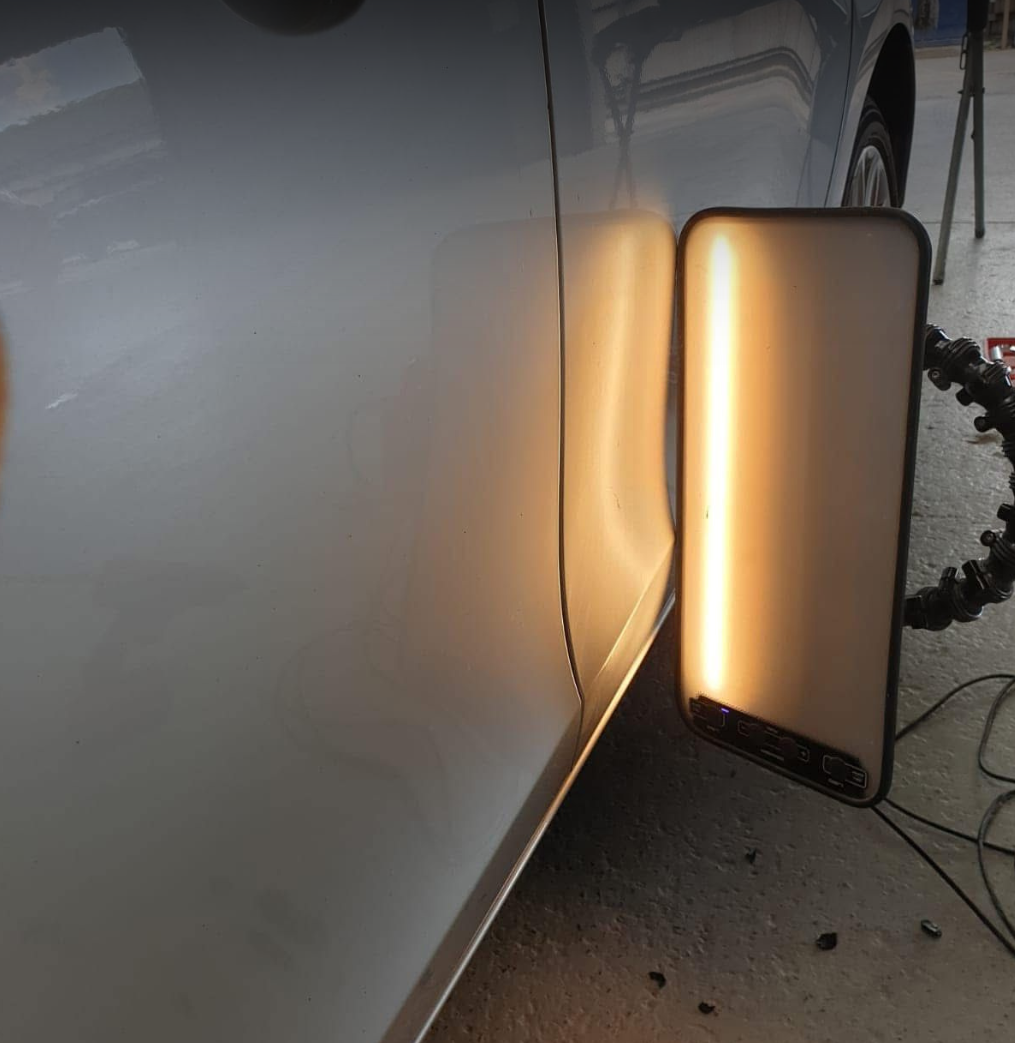 A close-up of a car's shiny grey body with a reflective surface, next to a lighted mirror or display on a stand, showing part of a garage or workshop environment.
