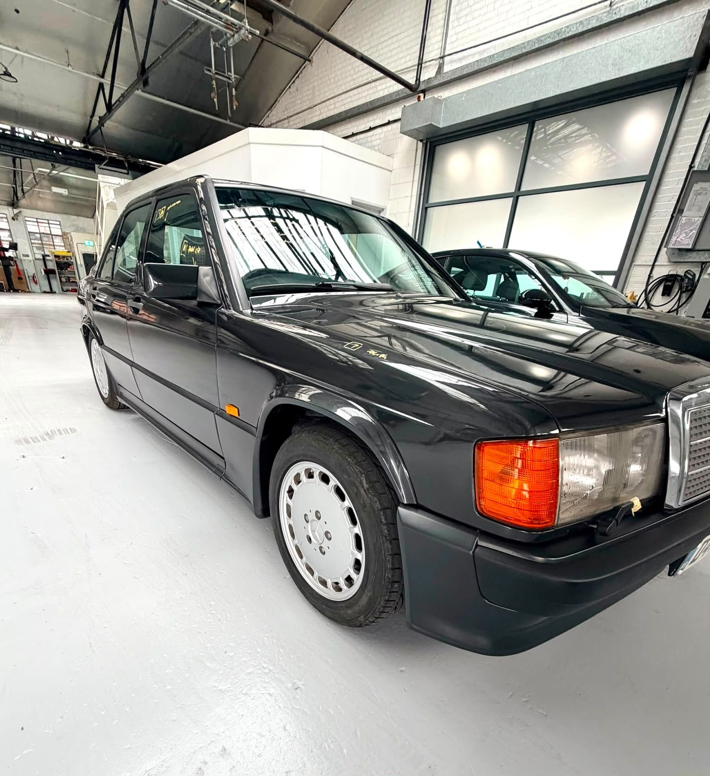 A black vintage Mercedes-Benz sedan parked inside a spacious industrial garage with large windows and white painted floors.