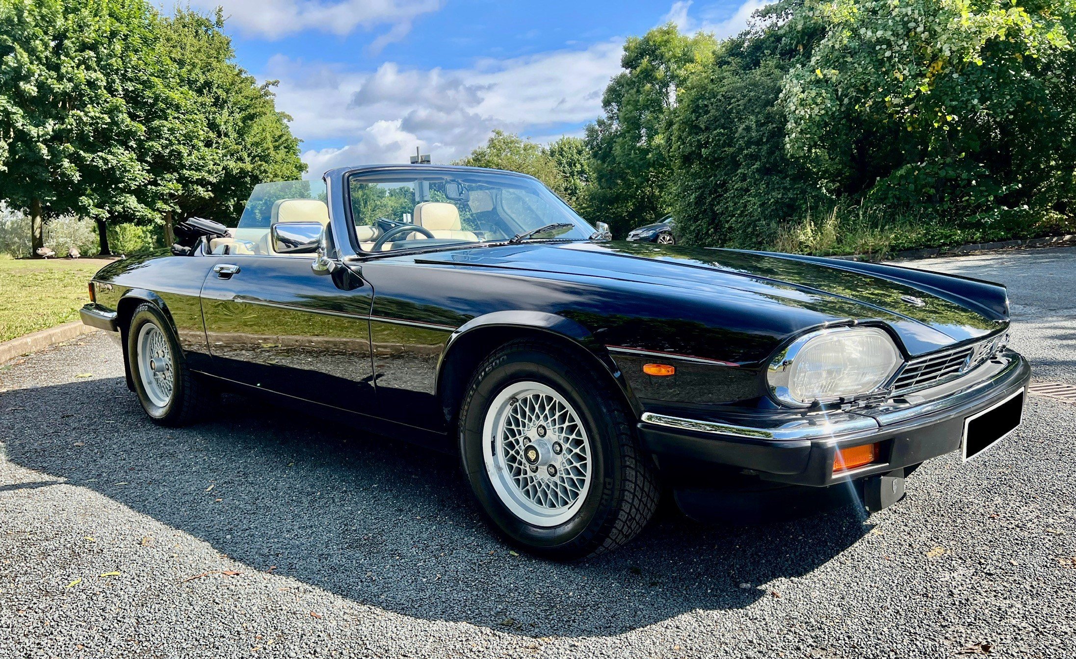 A black convertible sports car with tan interior parked outdoors on a gravel surface, with trees and blue sky in the background.