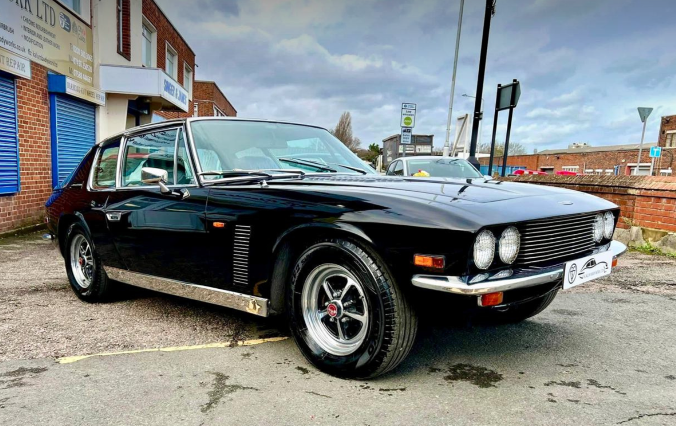 Black vintage sports car parked on a city street with brick buildings and a cloudy sky in the background.