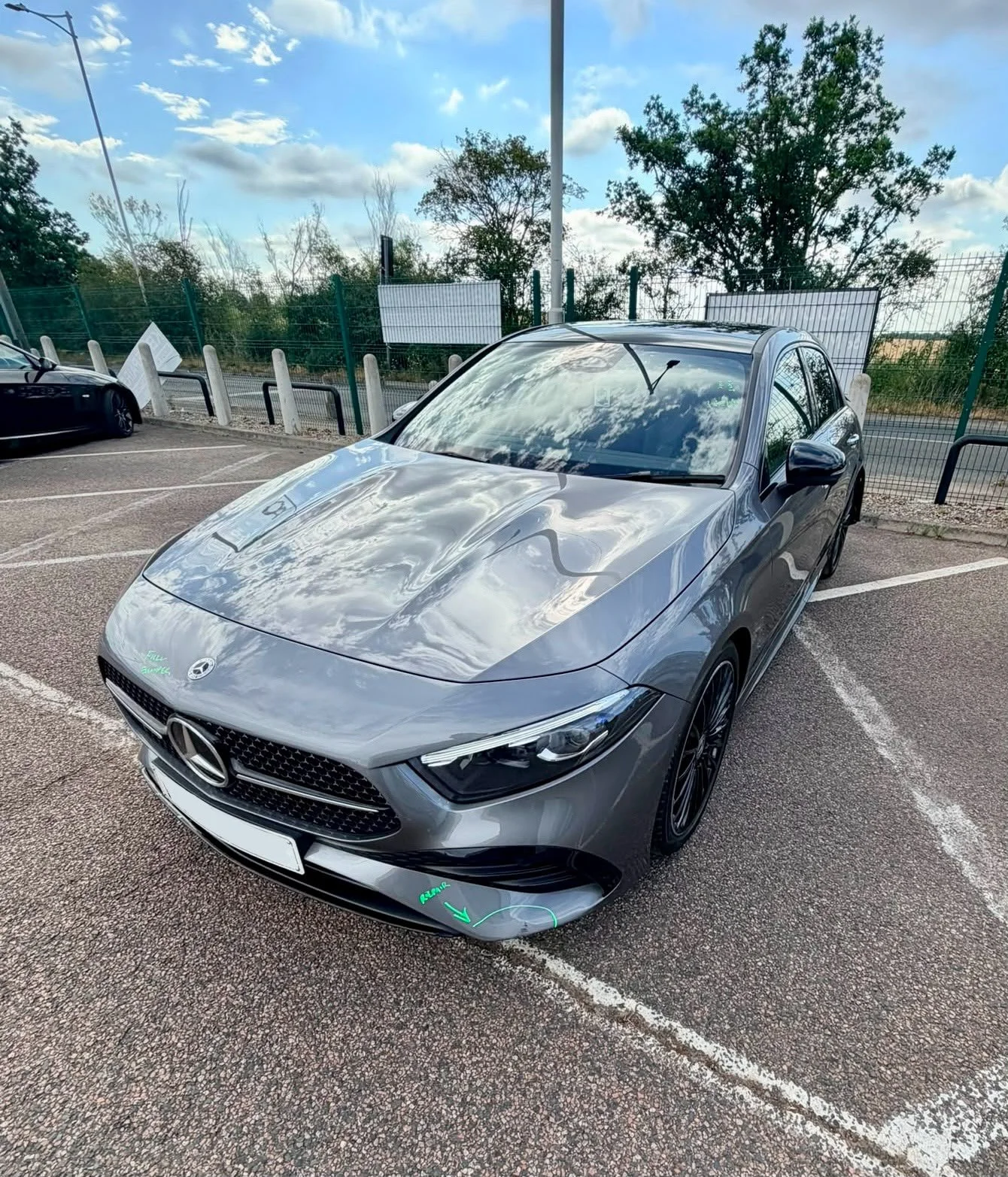 A silver Mercedes-Benz car parked in a parking lot with trees and a fence in the background. The sky is partly cloudy and the car's surface reflects the sky and clouds.