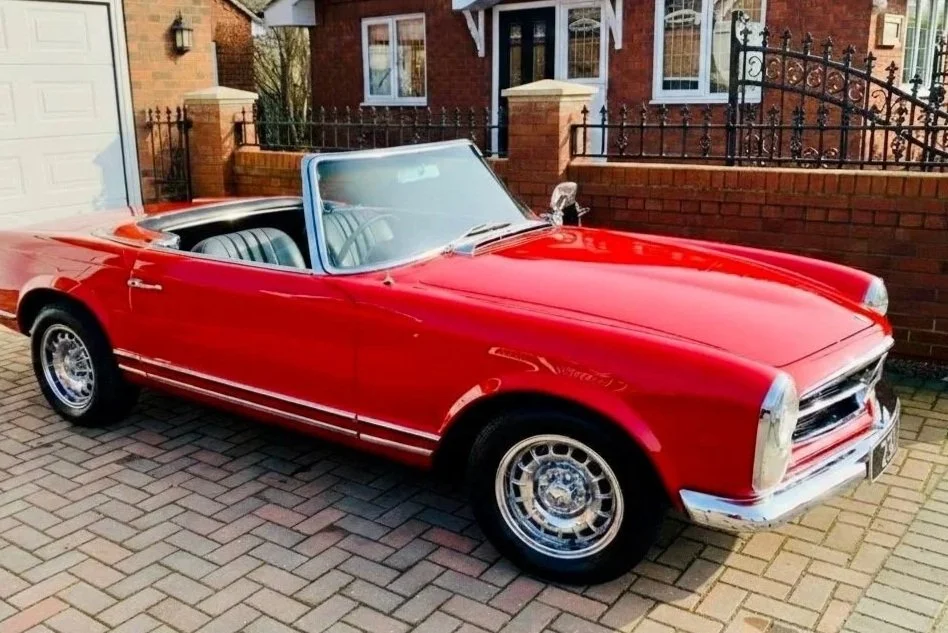 Red vintage convertible car parked on brick driveway in front of a brick house with white window trims and a black wrought iron fence.