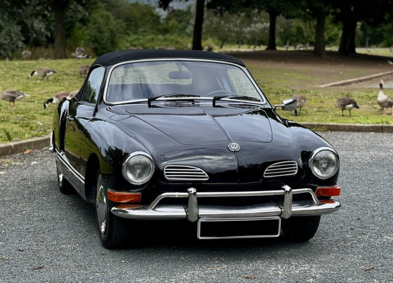 A black vintage Volkswagen Karmann Ghia car parked on a gravel surface with ducks and geese in a park-like setting in the background.