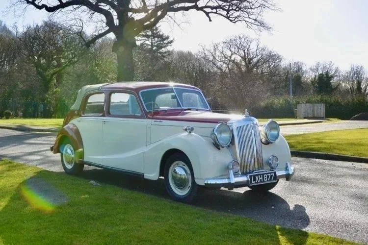 A vintage cream-colored car with red accents parked on a paved road beside a grassy area under a partly cloudy blue sky.