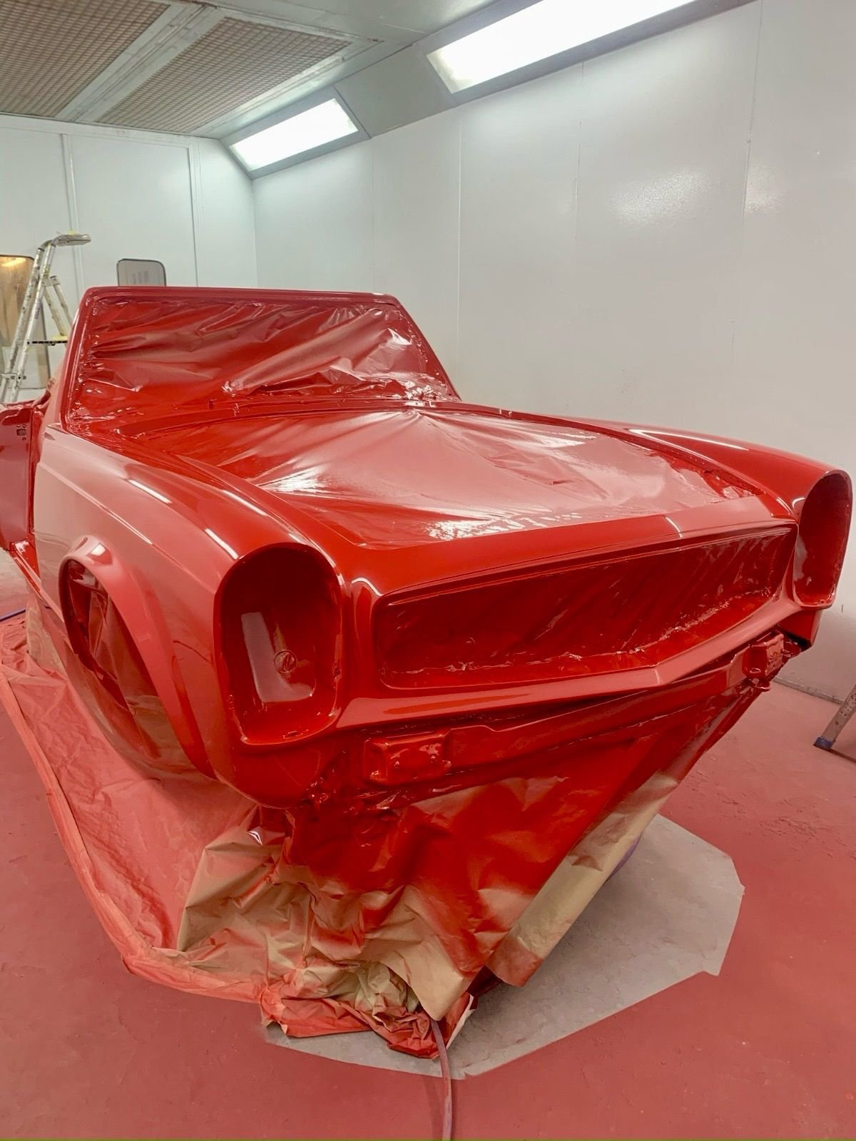 A red vintage car body shell being painted in a spray booth with white walls and ceiling lights.
