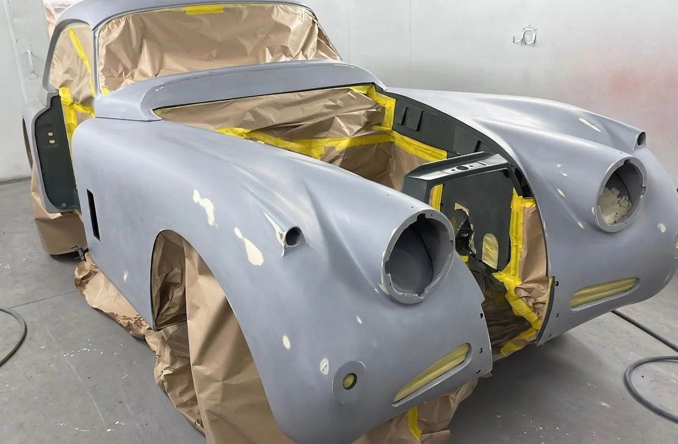 A classic car body shell in a paint booth, partially masked and primed, ready for painting.