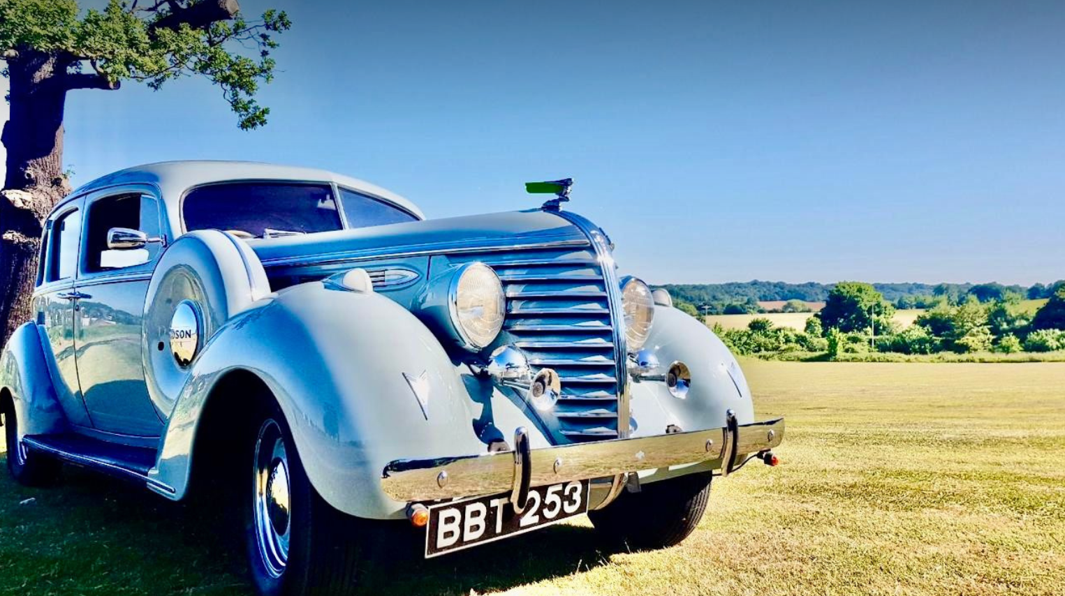 Vintage silver-blue car parked on grassy field with trees and blue sky in the background.