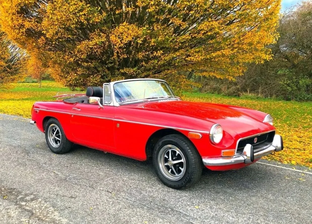 A vintage red convertible car parked on a gravel road with autumn trees and fallen leaves in the background.