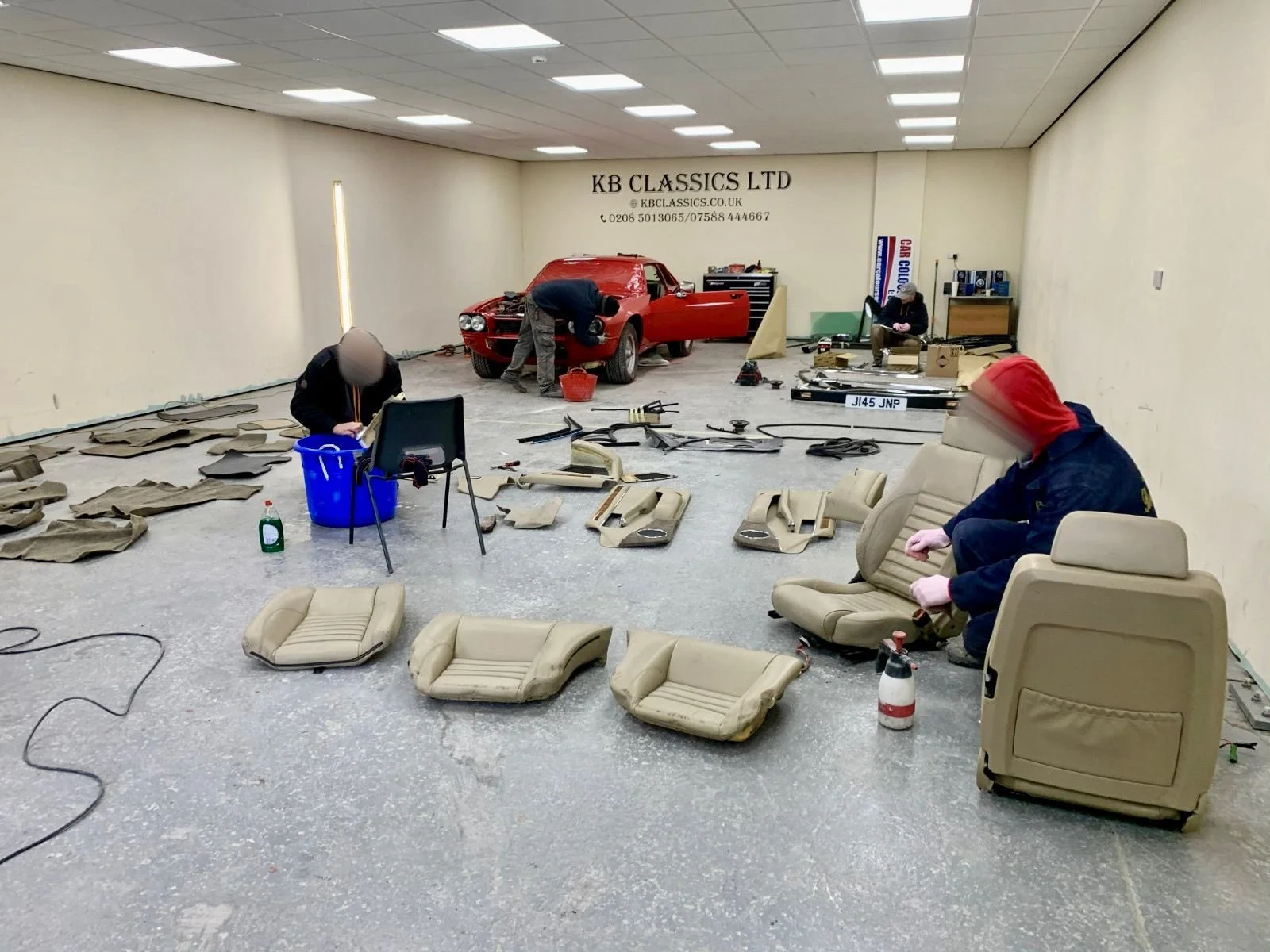 People working on a classic car in a large workshop, with disassembled car interior seats and tools scattered around.