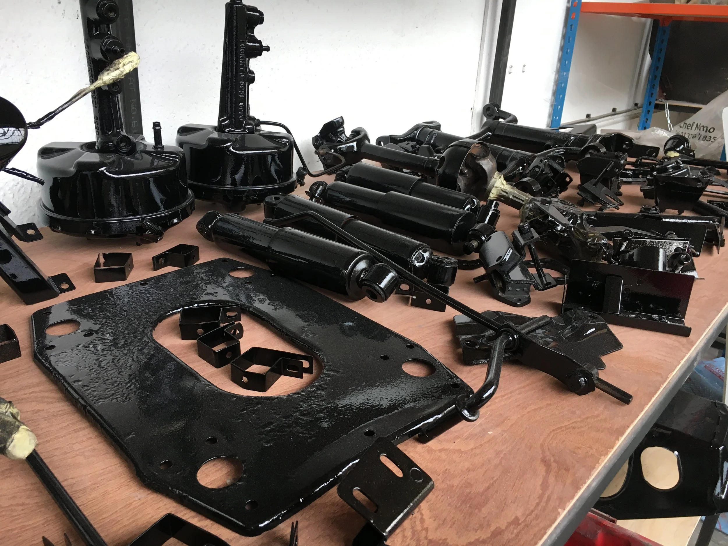 Various black mechanical parts and components laid out on a wooden table, with a blue and orange shelving unit in the background.