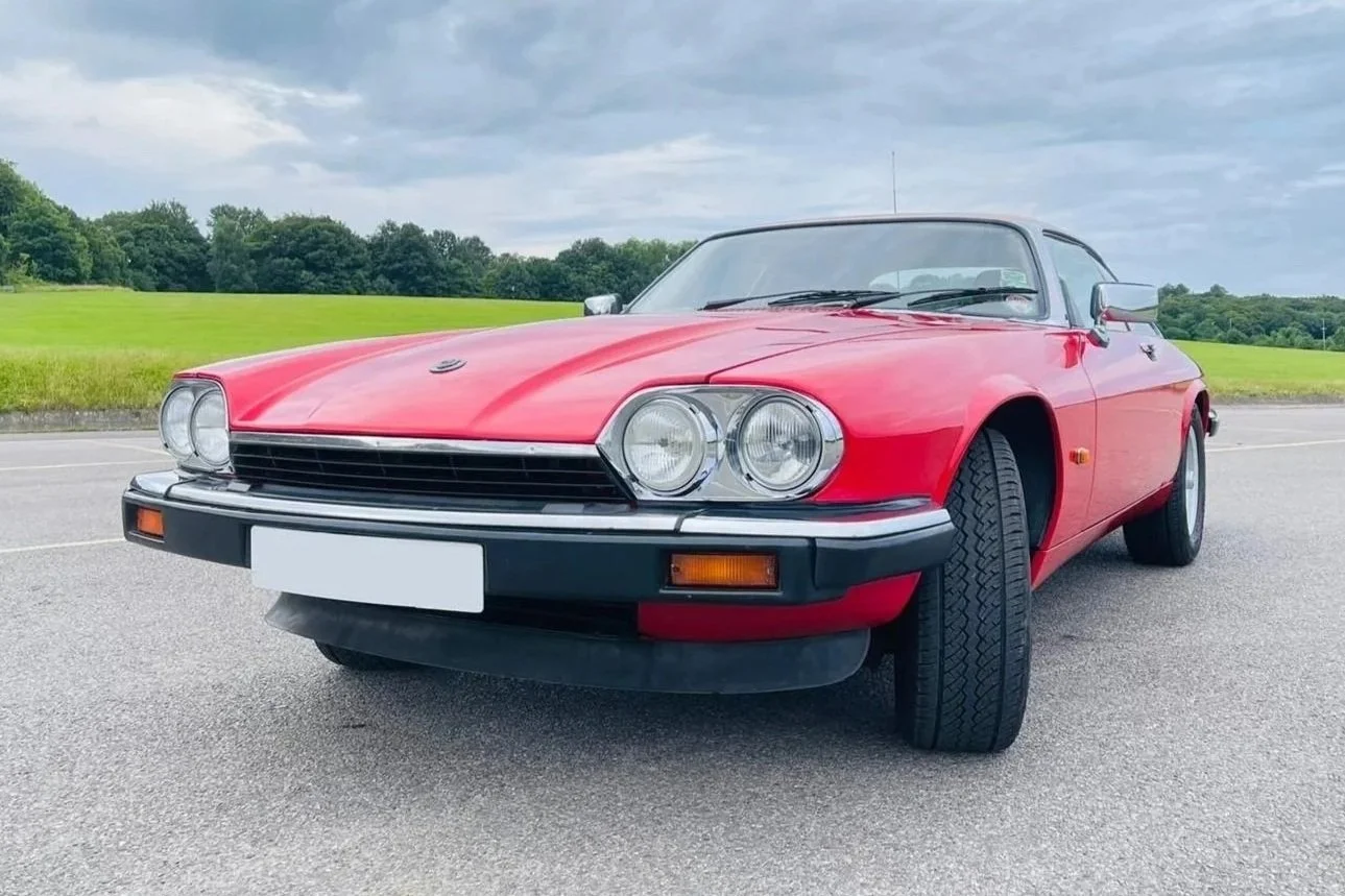 Red vintage sports car parked on a paved road with green grass and trees in the background under a cloudy sky.